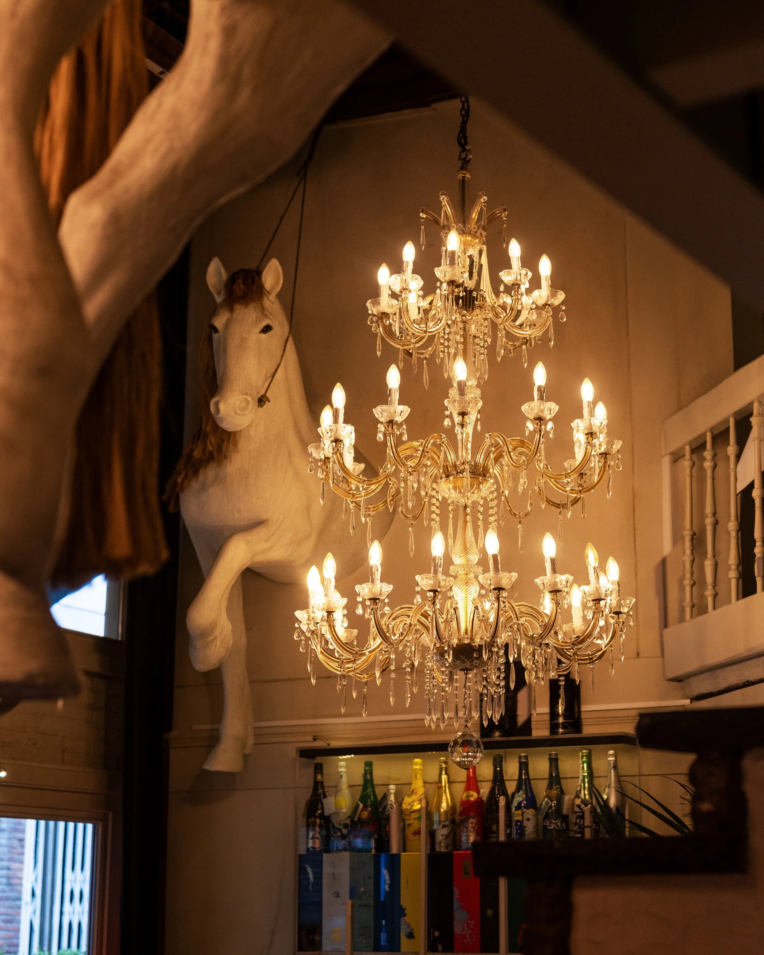An ornate chandelier with multiple candles hanging from the ceiling, next to a mounted horse head sculpture in a warmly lit interior.