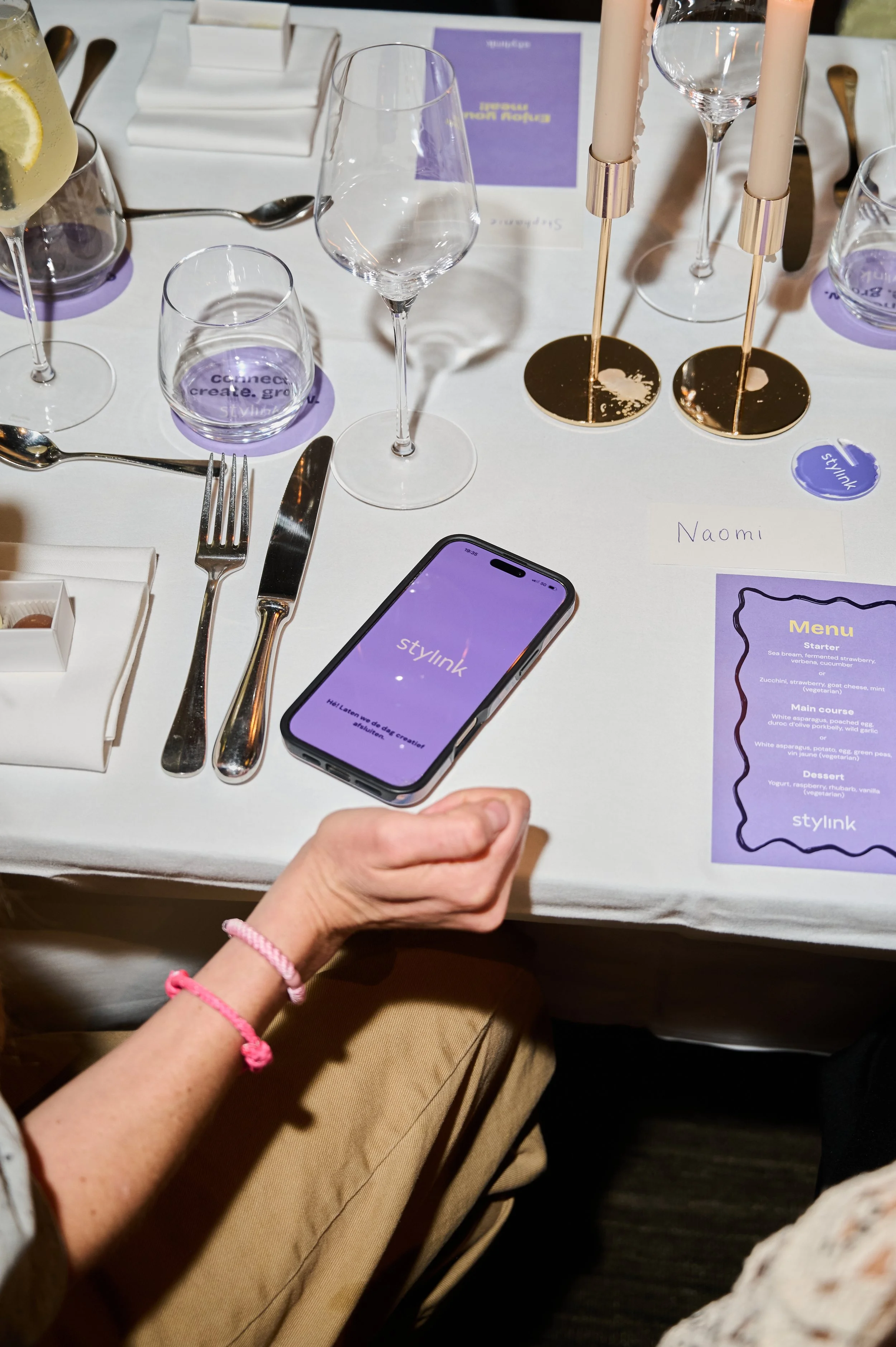 A person holding a smartphone with the StyLink app open, sitting at a table set for a meal with wine glasses, candles, and menus. The table has purple accent decor, and there are other glasses and utensils present.
