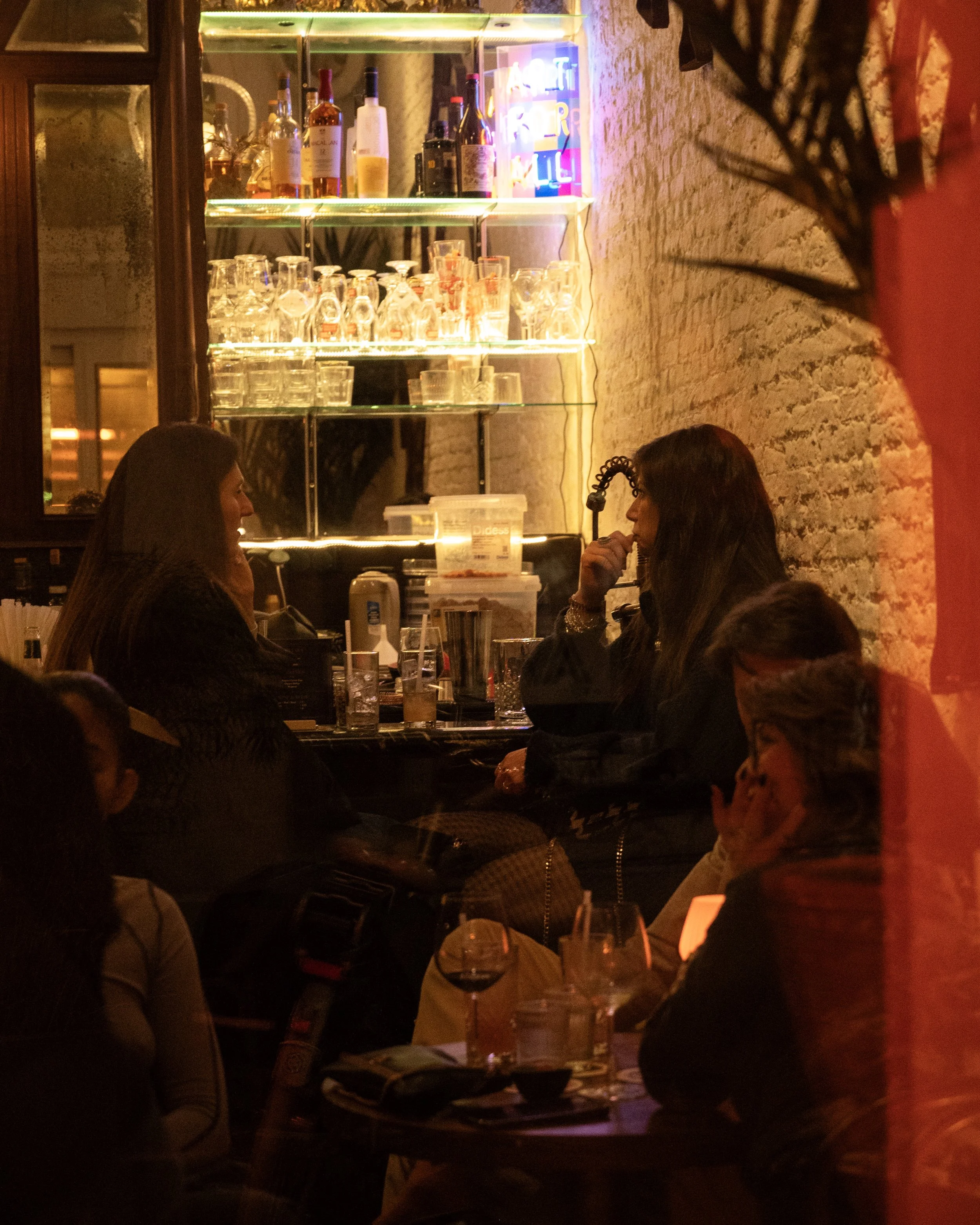 A dimly lit bar scene with three women sitting and talking, with glasses and drinks on the tables. The bar shelves in the background hold various bottles and glasses, and there is a neon sign on the wall.