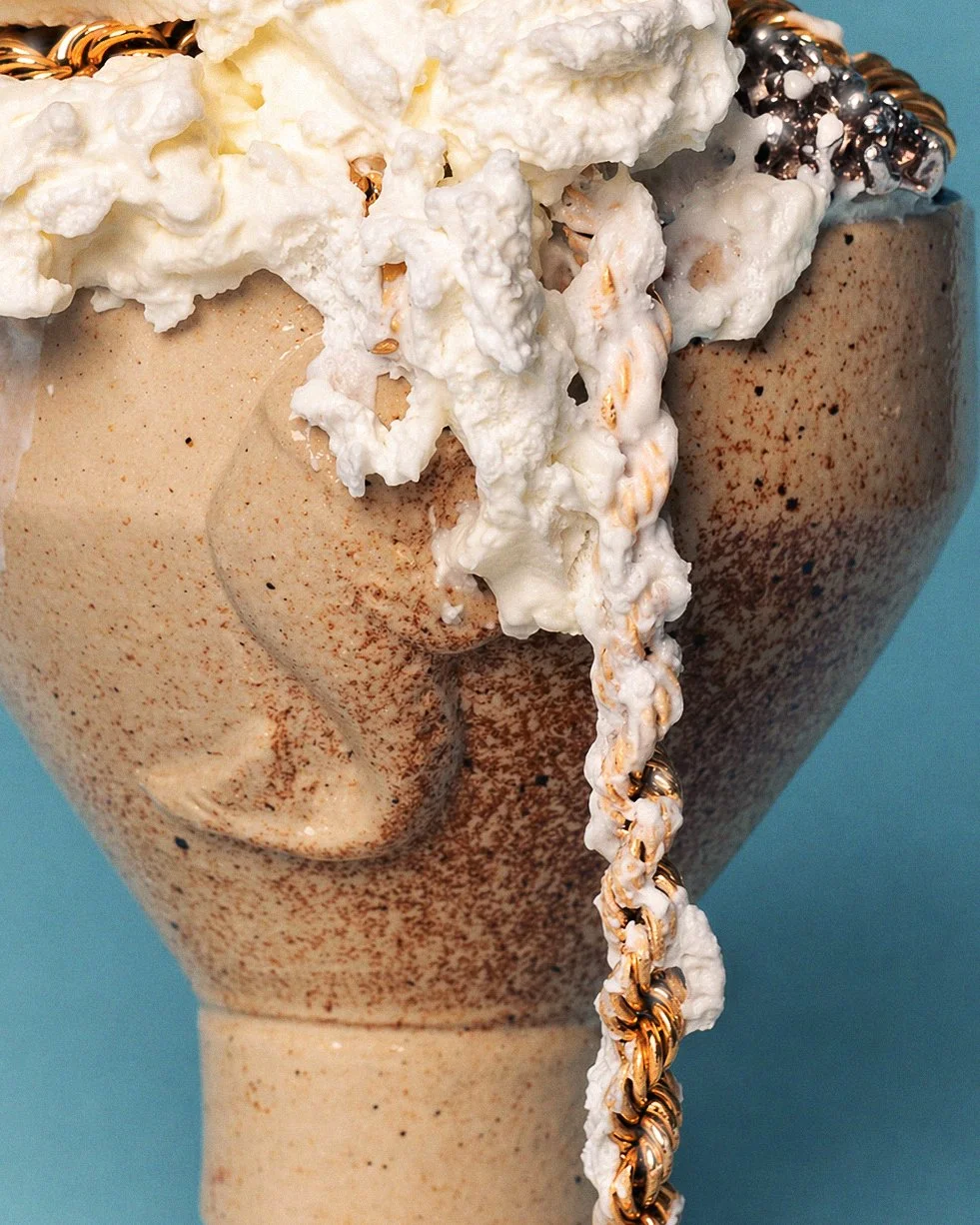 Close-up of an ice cream sundae in a beige speckled ceramic cup, topped with whipped cream, a chocolate and caramel topping, and a gold beaded bracelet or chain hanging from the whipped cream. The background is a plain teal color.