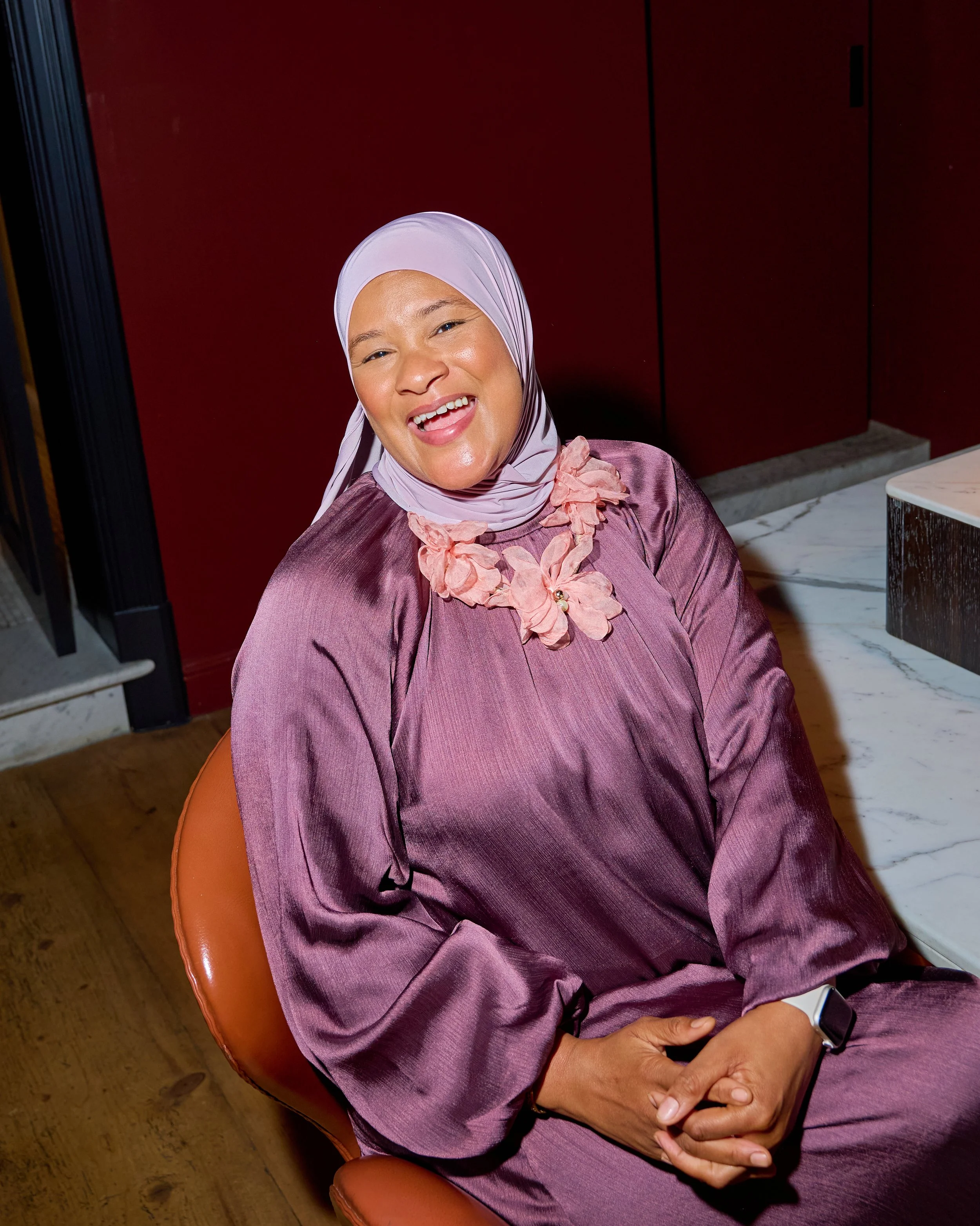 A woman wearing a light purple hijab and a shiny purple dress sitting on an orange chair with a pink floral necklace, smiling and holding her hands together, with a red wall behind her.