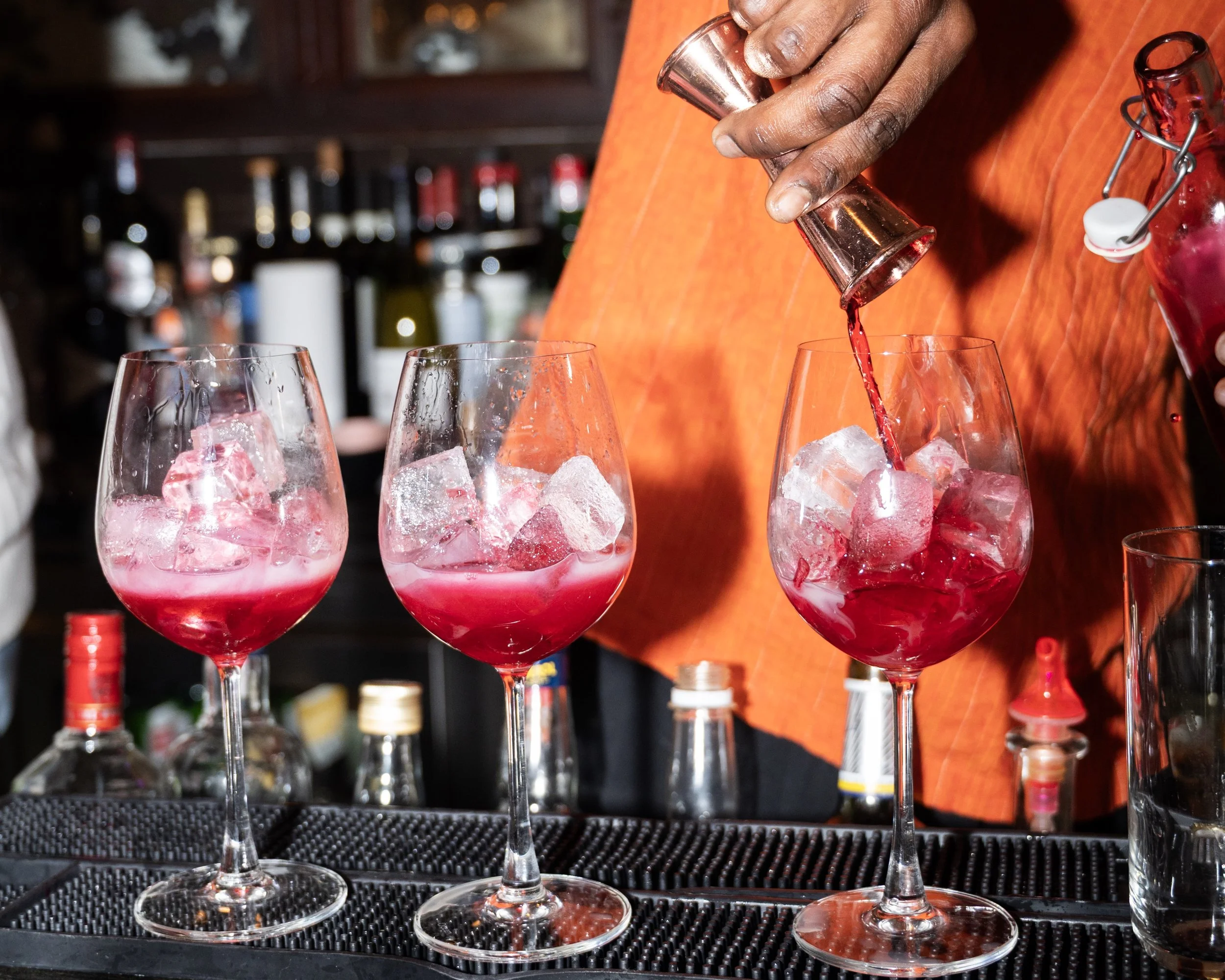 Bartender pouring red cocktail into large wine glass filled with ice at bar counter.
