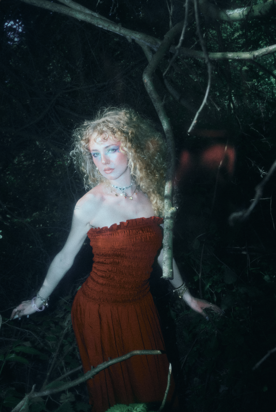A woman with curly blonde hair, wearing a red dress and jewelry, standing among trees and branches in a dark forest.