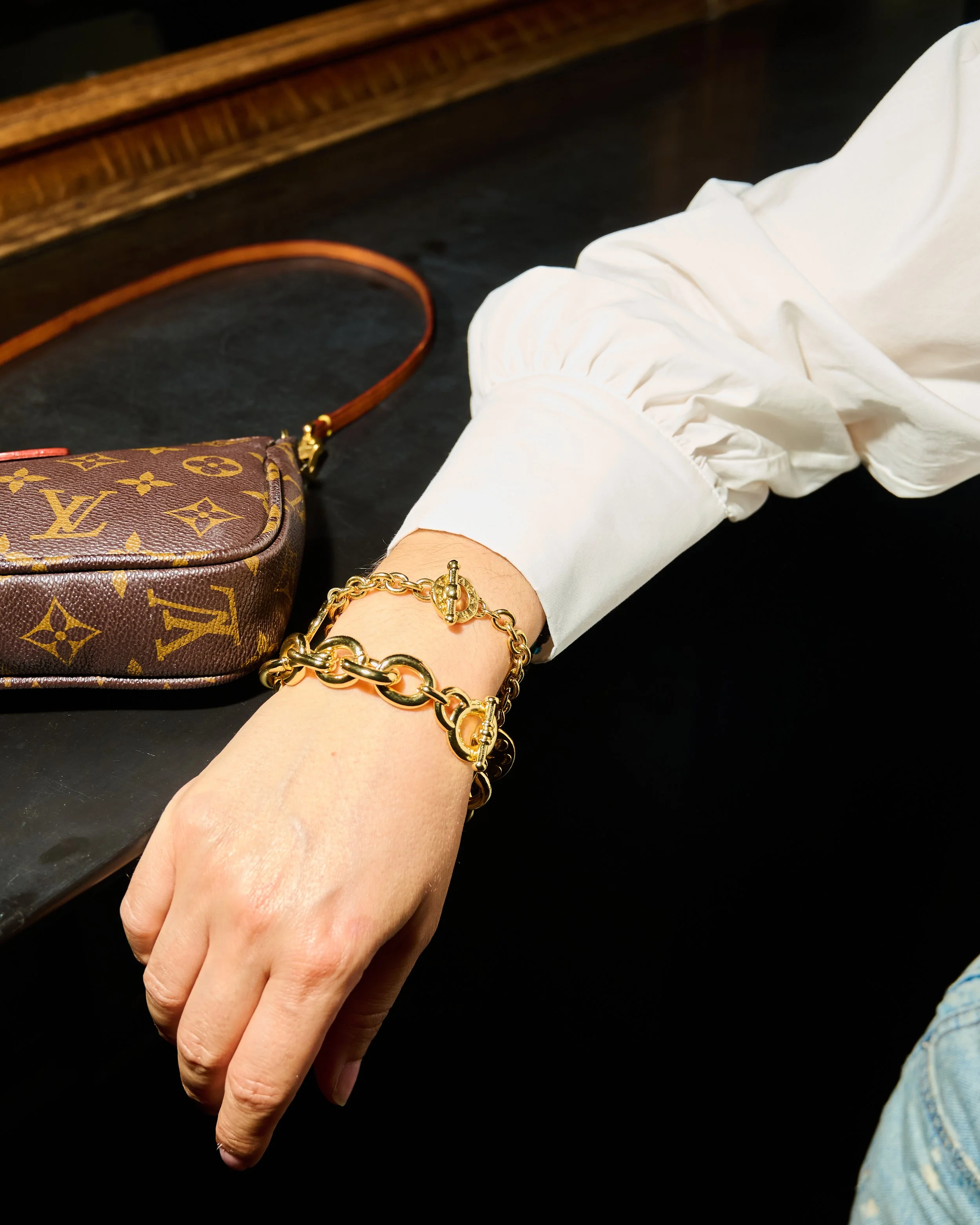 A hand wearing gold jewelry resting on a dark surface, with a Louis Vuitton monogram bag and a white long sleeve shirt visible in the background.