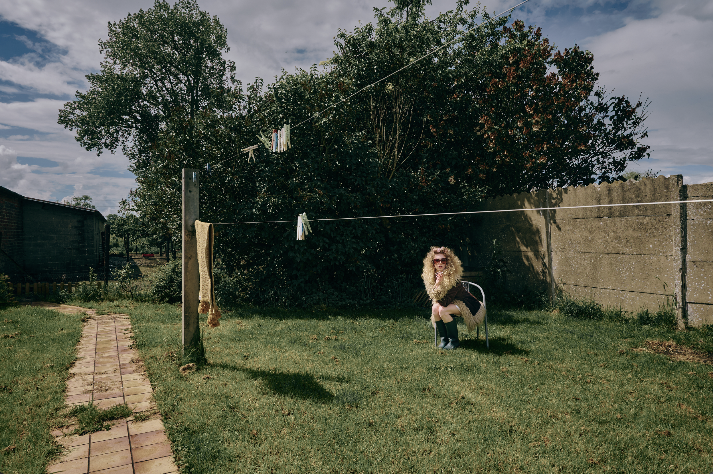 A woman with curly blonde hair, wearing sunglasses and a fur-trimmed black jacket, sitting on a white chair in a backyard, with clothes hanging on a clothesline behind her. The yard has a brick pathway, green grass, and trees, under a partially cloud