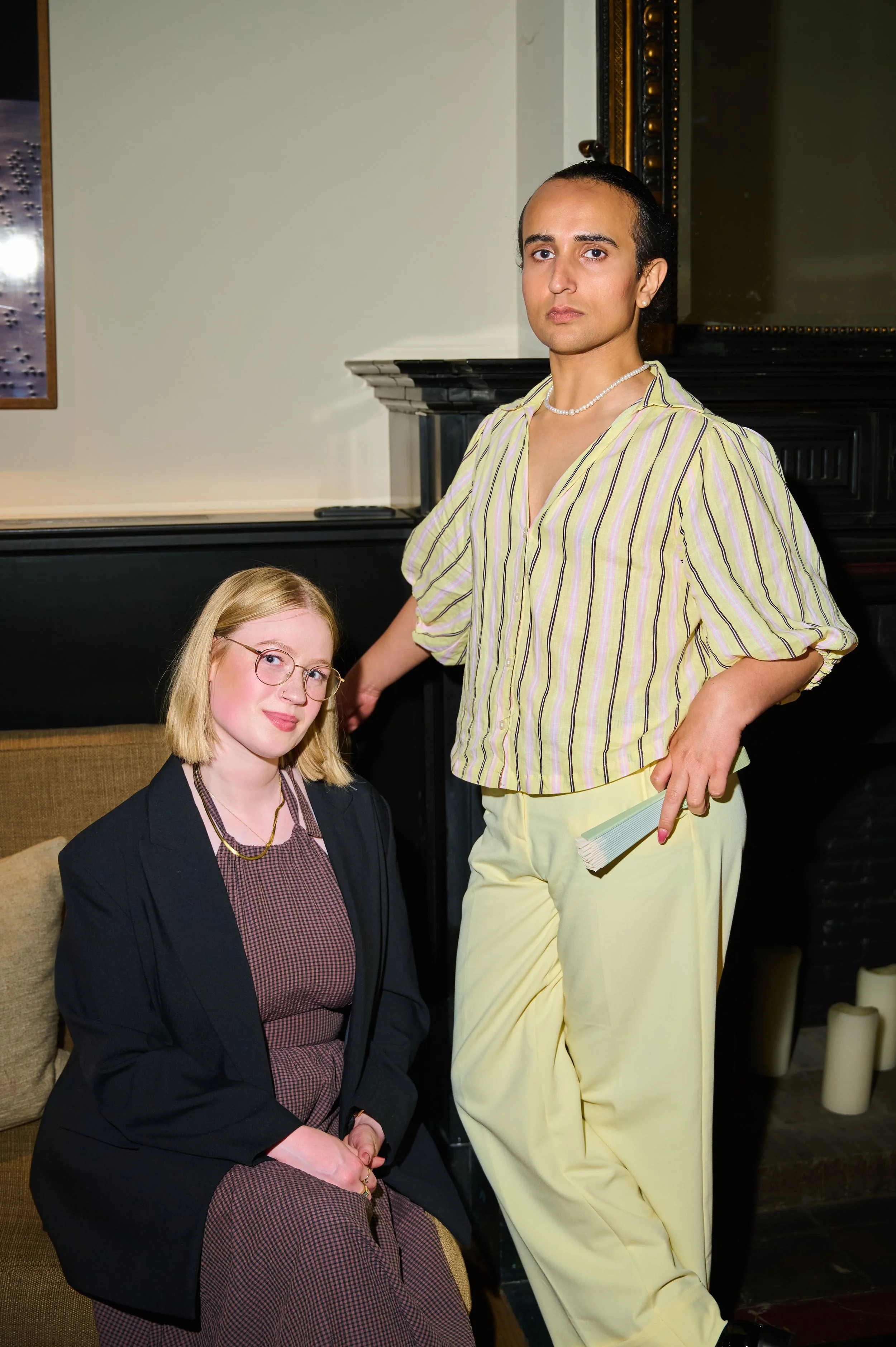 A man and a woman pose together indoors, with candles on the floor, a black piano, and a dark ornate mirror in the background. The man is standing, wearing a yellow striped shirt and yellow pants, holding a book, and the woman is sitting, wearing a d