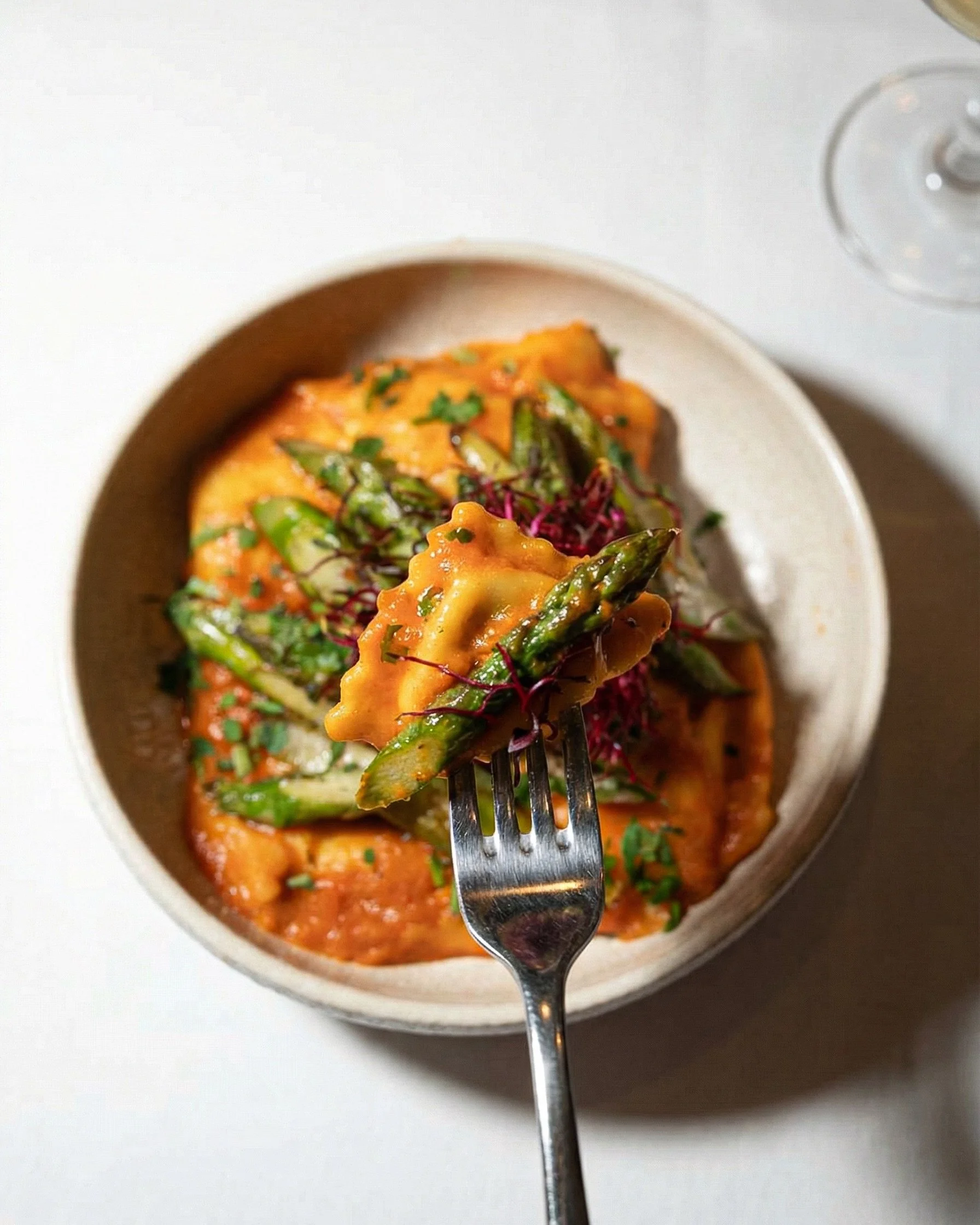 A fork holding a bite of ravioli with green asparagus and red microgreens over a bowl of ravioli with tomato sauce, garnished with chopped herbs.