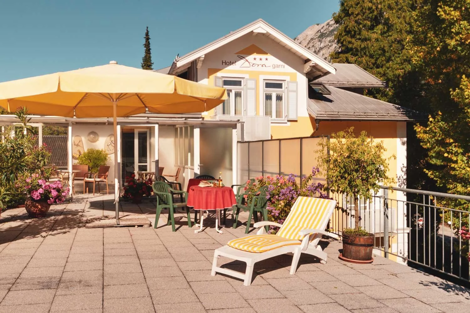 Terrasse vor einem gelben Hotel mit Tisch, Stühlen, Sonnenschirm und Pflanzen, bei sonnigem Wetter