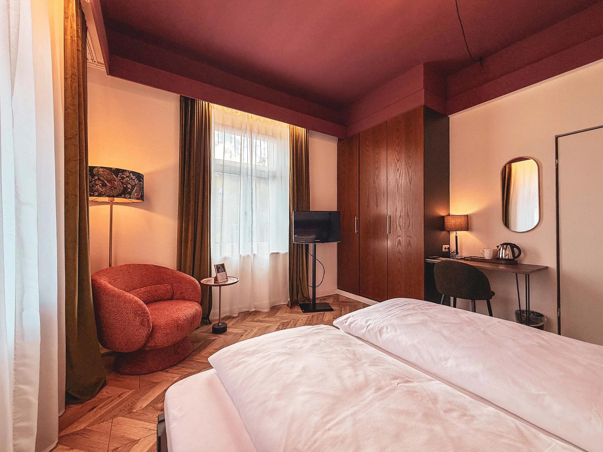 Ein stilvoll eingerichtetes Hotelzimmer mit beigem Bett, rotem Ohrensessel, einem kleinen runden Tisch, Fernseher, einem Holzschreibtisch mit Lampe, Büchern, einem Wasserkocher und einem Spiegel. Die Fenster sind mit weißen und grünen Vorhängen dekoriert.