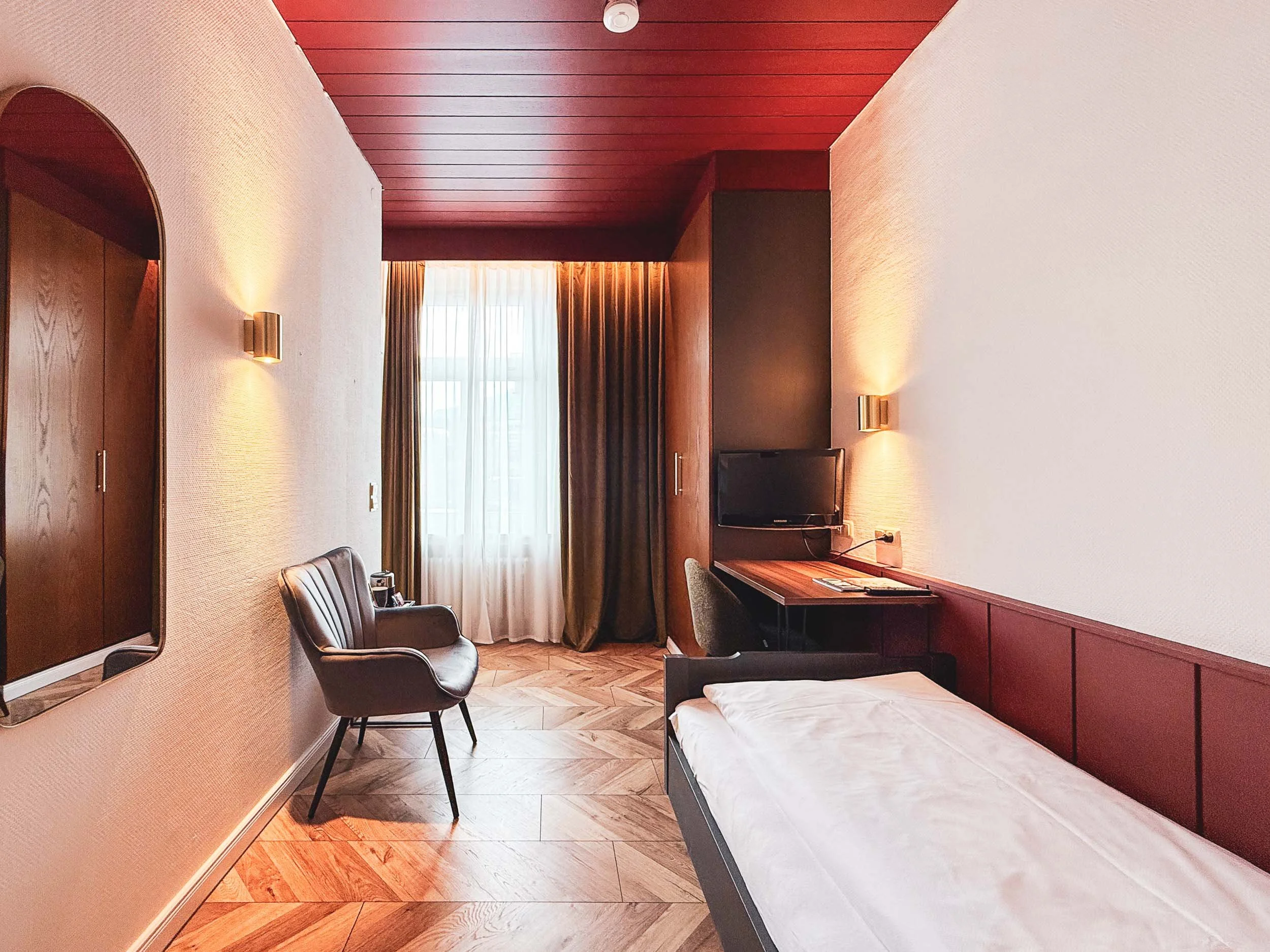 Ein modernes Hotelzimmer mit Einzelbett, Fernseher am Schreibtisch, einem Sessel und einem Fenster mit Vorhängen.