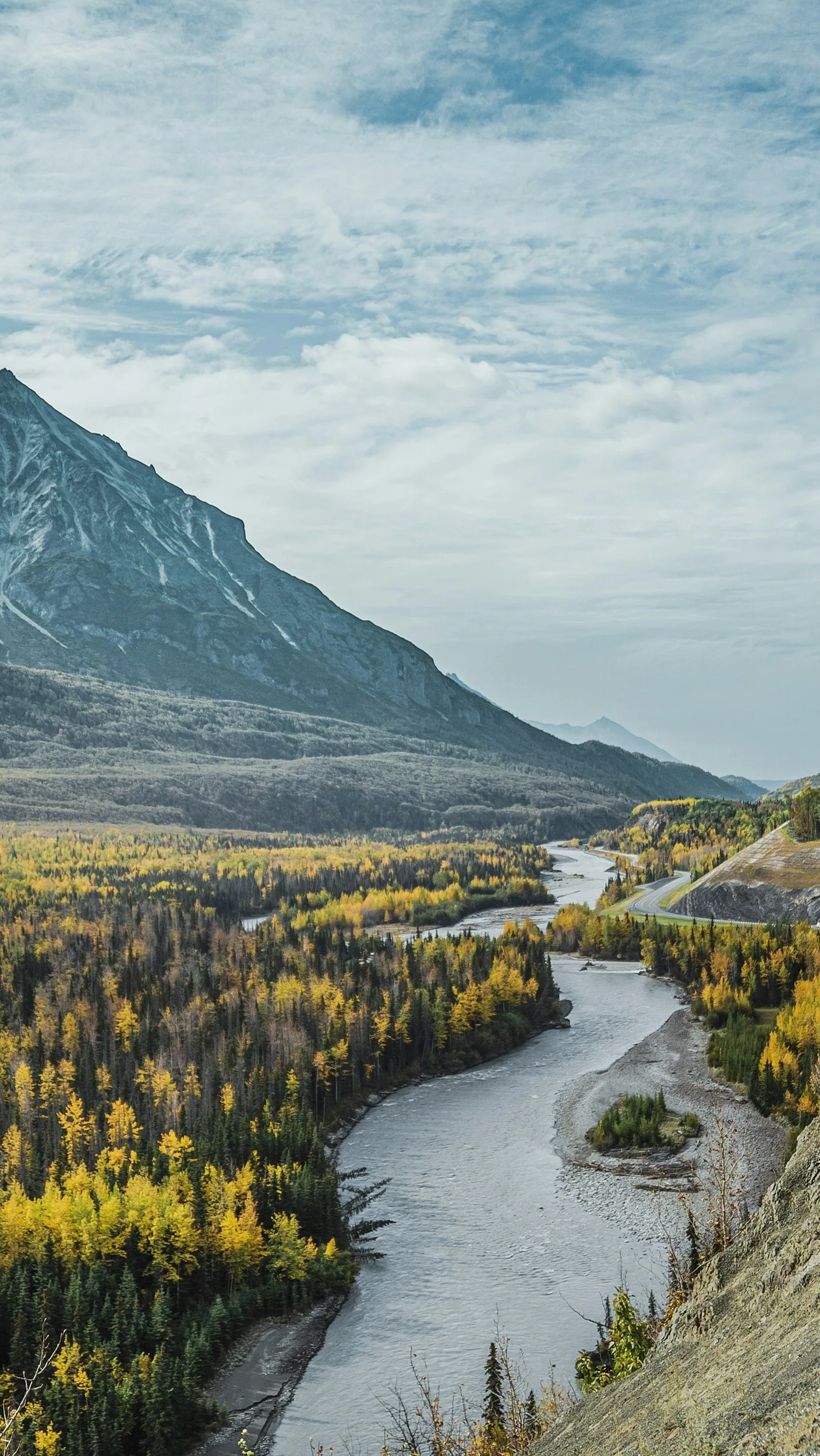 A scenic mountain landscape with a winding river through a forest with yellow and green trees, mountains in the background, and a partly cloudy sky.