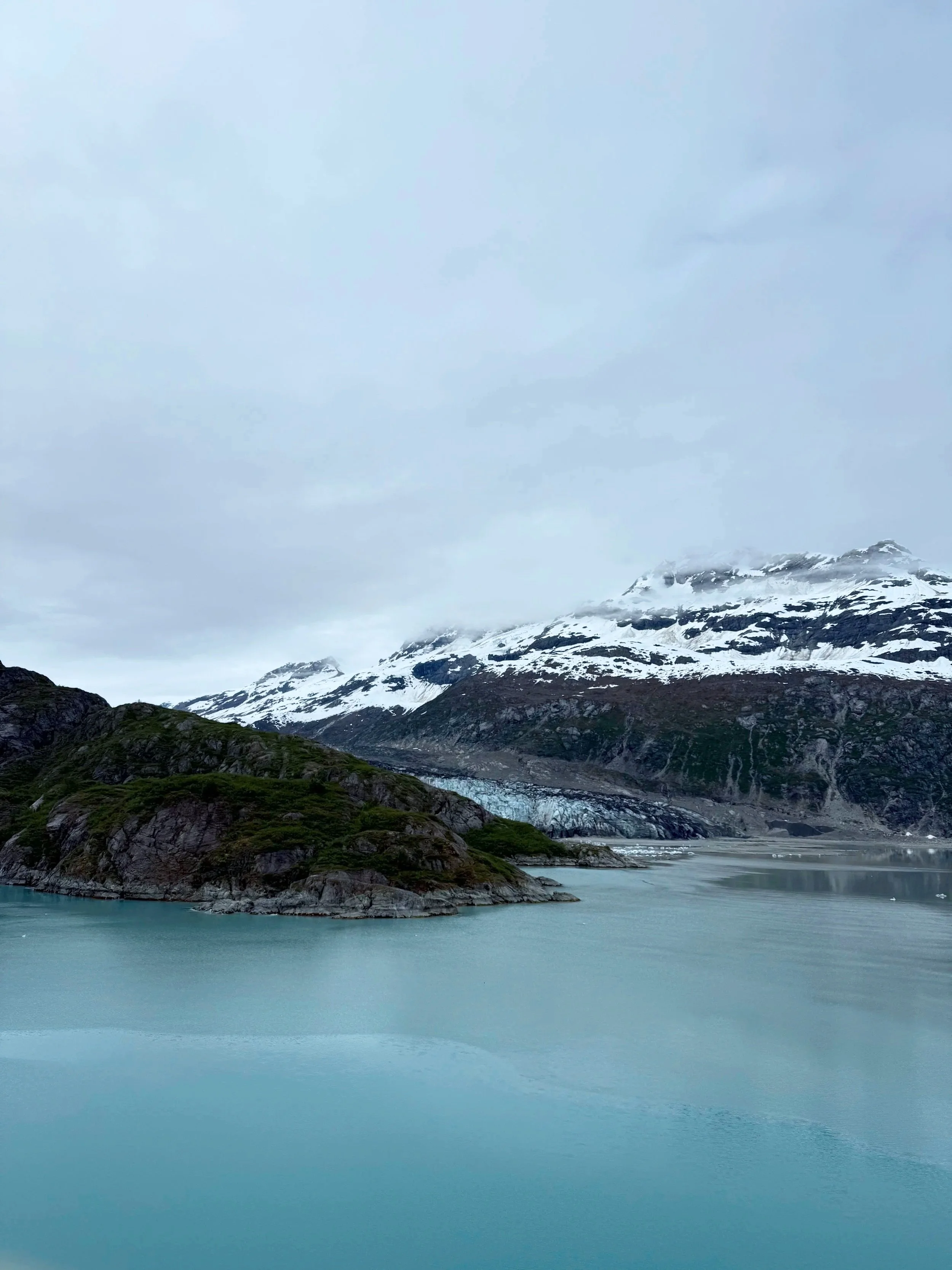 Snow-capped mountains above a calm turquoise glacier-fed body of water under a cloudy sky.