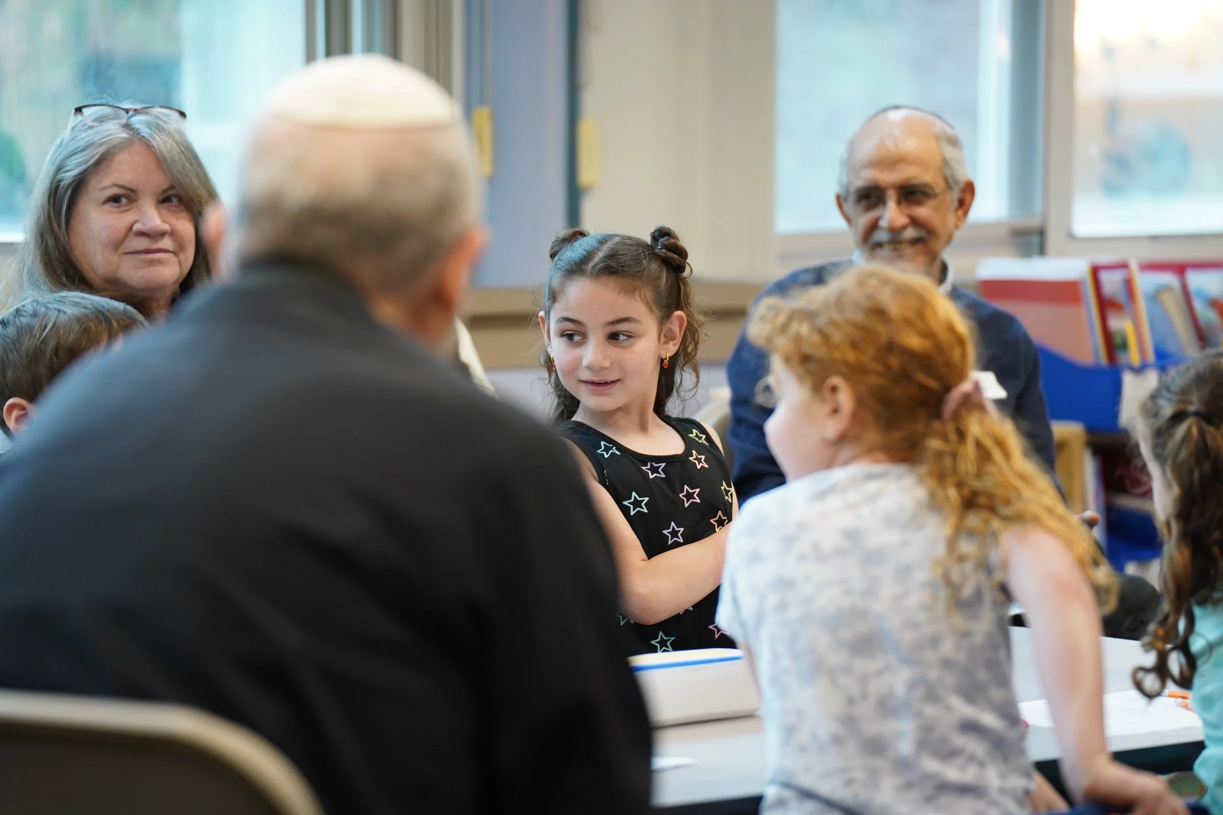 A young girl with dark hair in a bun and wearing a black dress with colorful stars is speaking to an older man and a woman in a classroom setting, with other children and adults present.