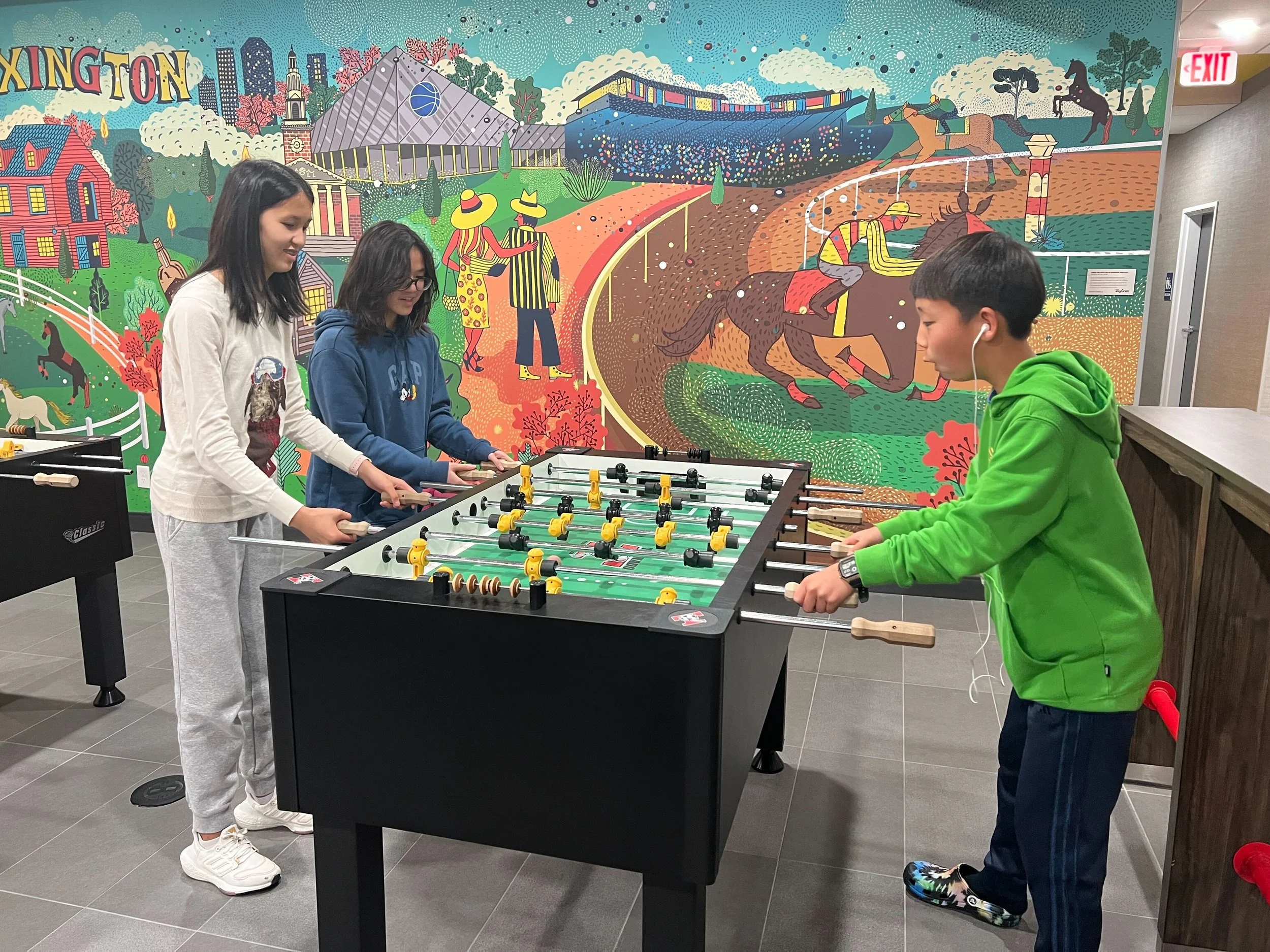 Three children playing foosball in an indoor recreation area with a colorful mural of a horse race and cityscape in the background.