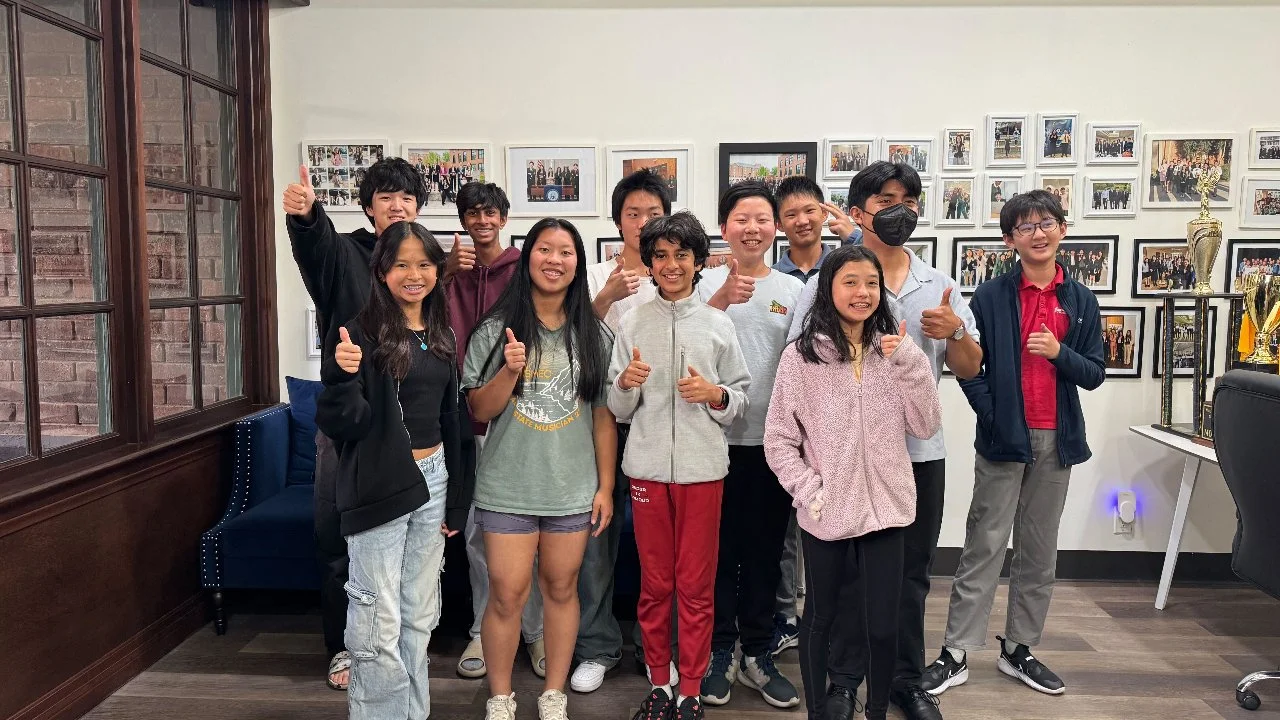 Group of ten diverse young people smiling and giving thumbs-up in an indoor room with photo walls and trophies.