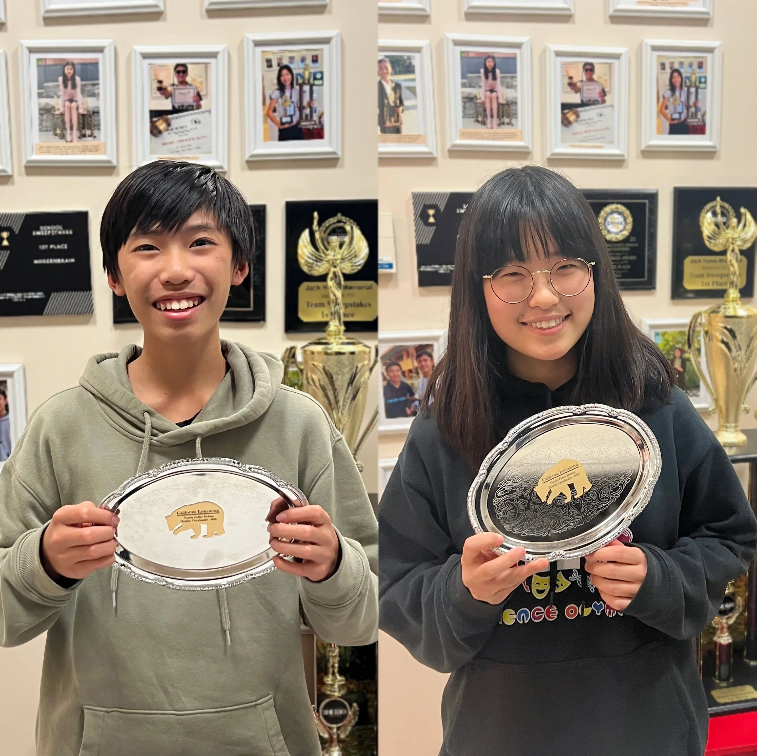 Two young people holding silver trays with a gold emblem of a bear. They are smiling and standing in front of a wall with trophies and framed photographs.