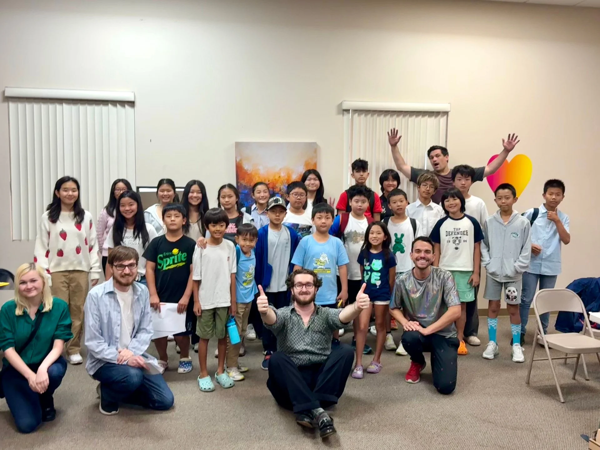 Group of smiling children and three adults posing together indoors, some giving thumbs up, with colorful decorations and artwork in the background.