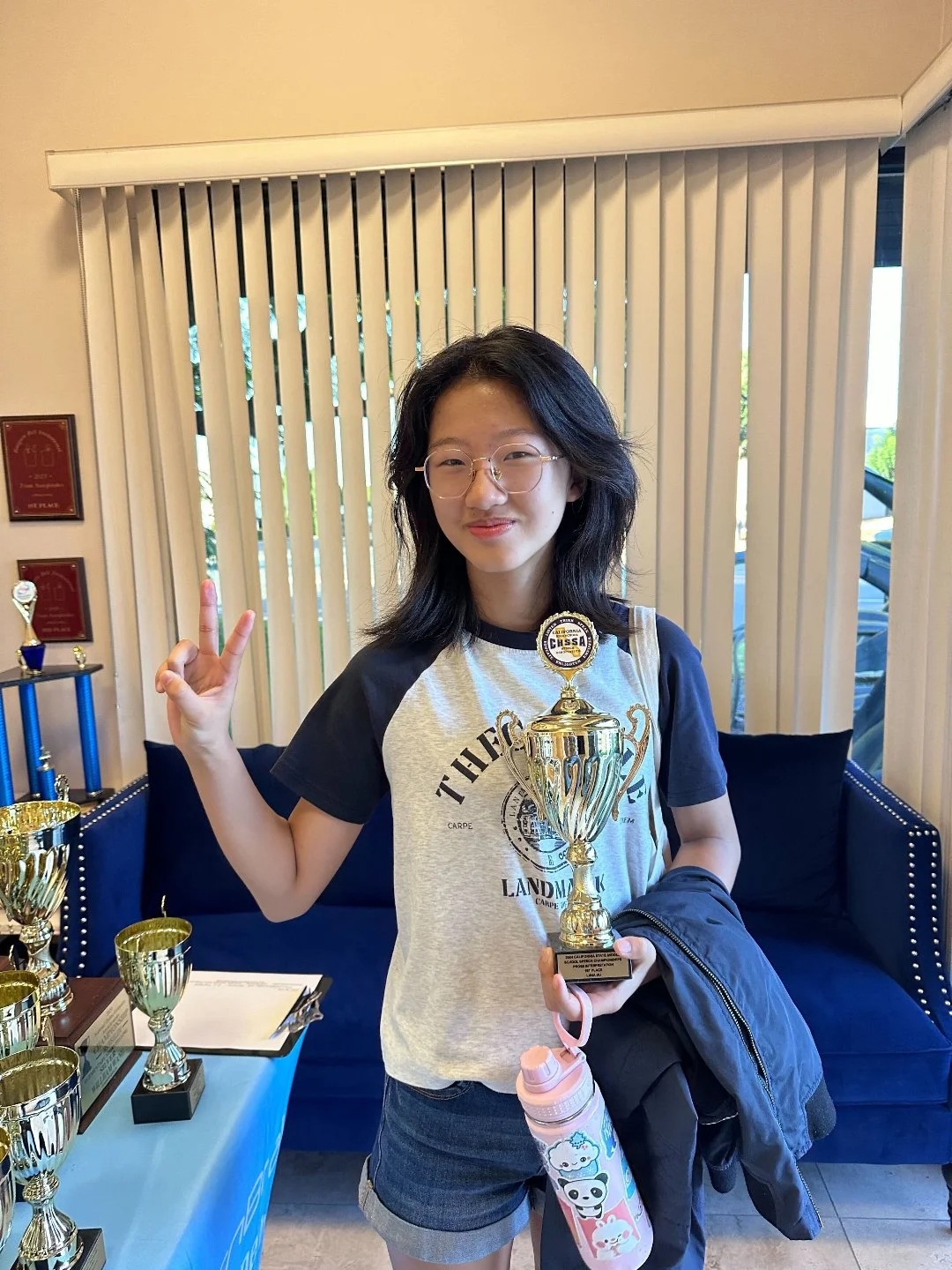 A young woman with glasses holding a large trophy and making a peace sign in a room with several other trophies on a table.