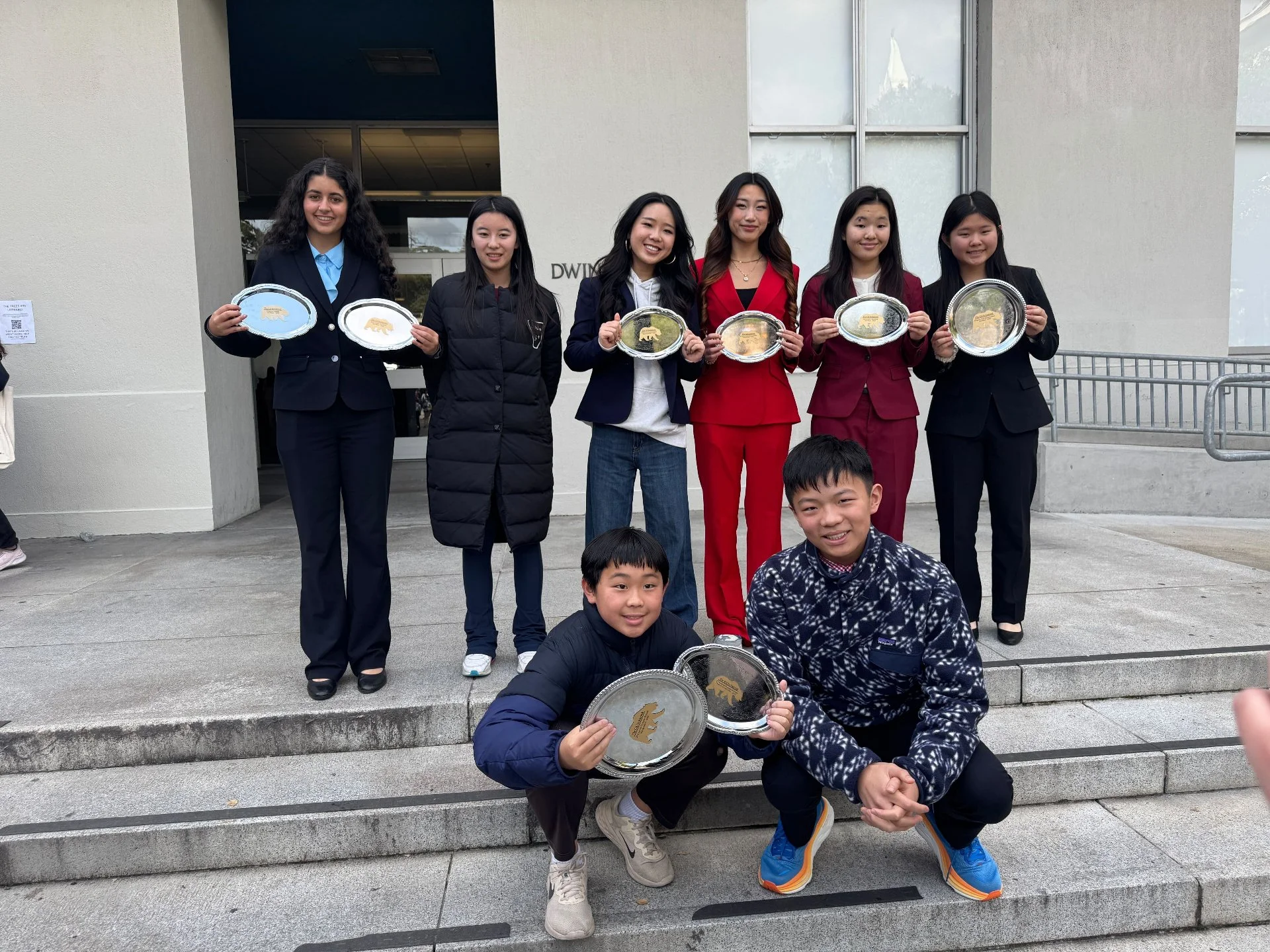 Group of eight students standing outside, holding silver award trays, two students crouching in front with trays, smiling after a competition or award ceremony.
