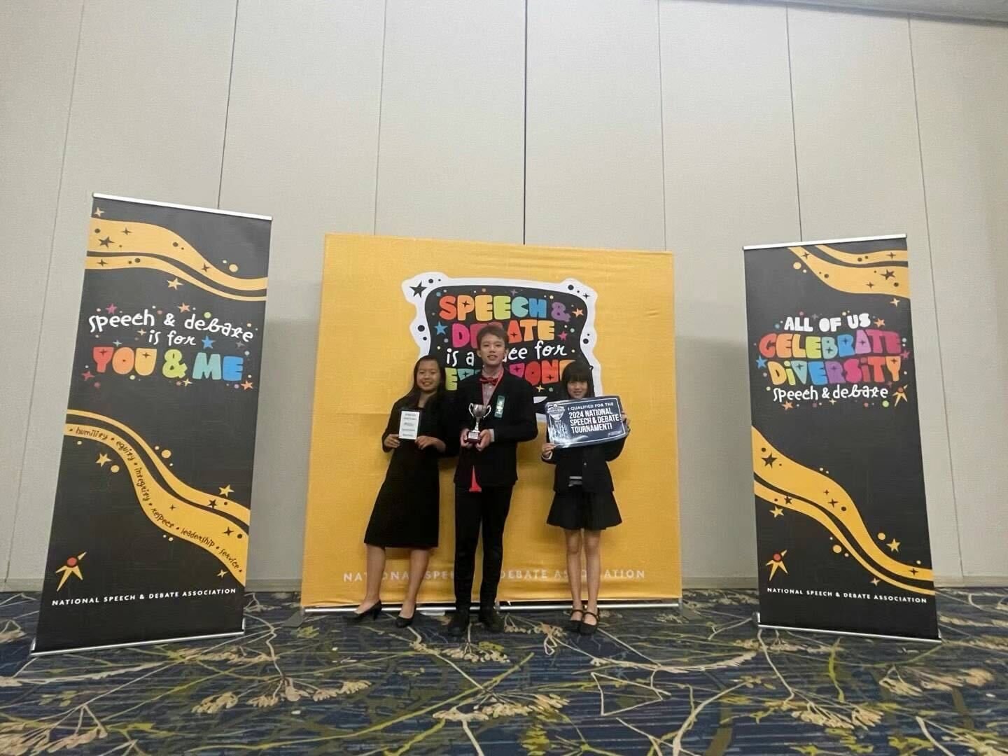Three young students standing on a stage at a speech and debate competition, holding awards, with colorful banners and a yellow backdrop behind them.