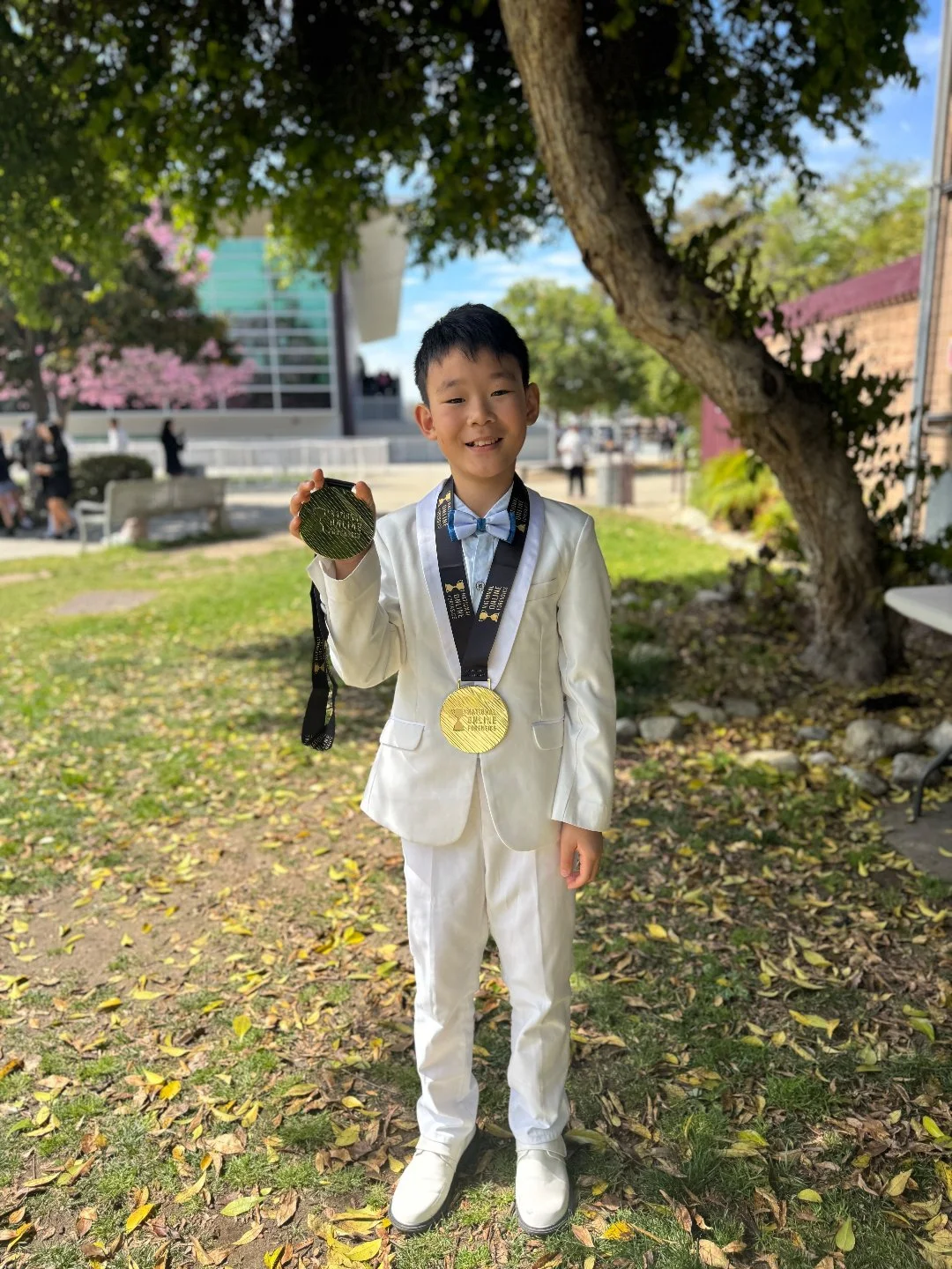 A young boy in a white suit holding a gold medal, standing outdoors under a large tree, smiling, with a building and people in the background.