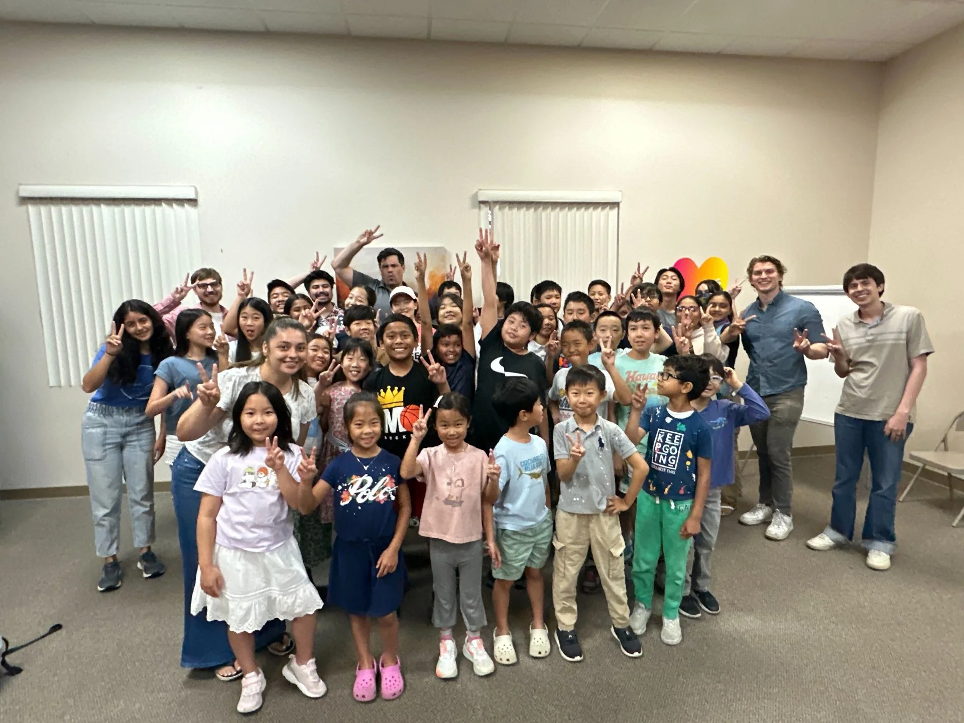 A large group of children and a few adults posing together indoors, making peace signs and smiling for the camera.