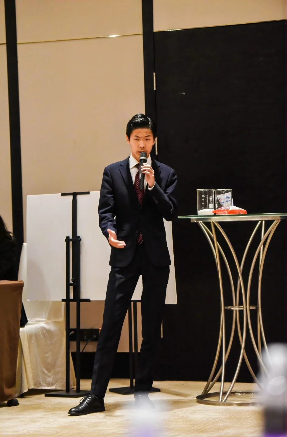 A young man in a dark suit, white shirt, and red tie is speaking into a microphone at a formal event, standing beside a small round table with a glass pitcher, a measuring cup, and some papers.