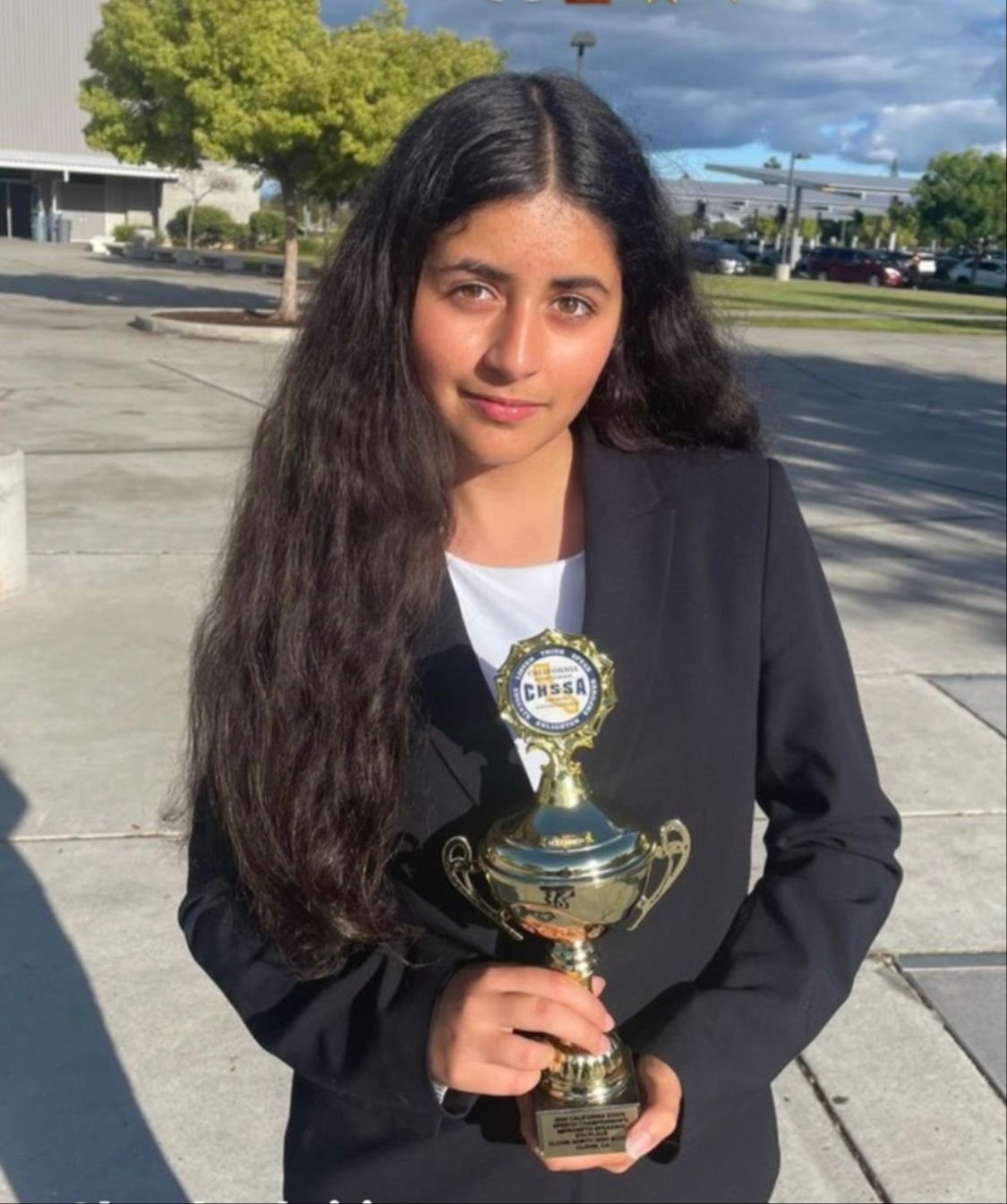 A young girl with long dark hair wearing a black blazer holds a championship trophy outdoors on a sunny day with trees and a parking lot in the background.