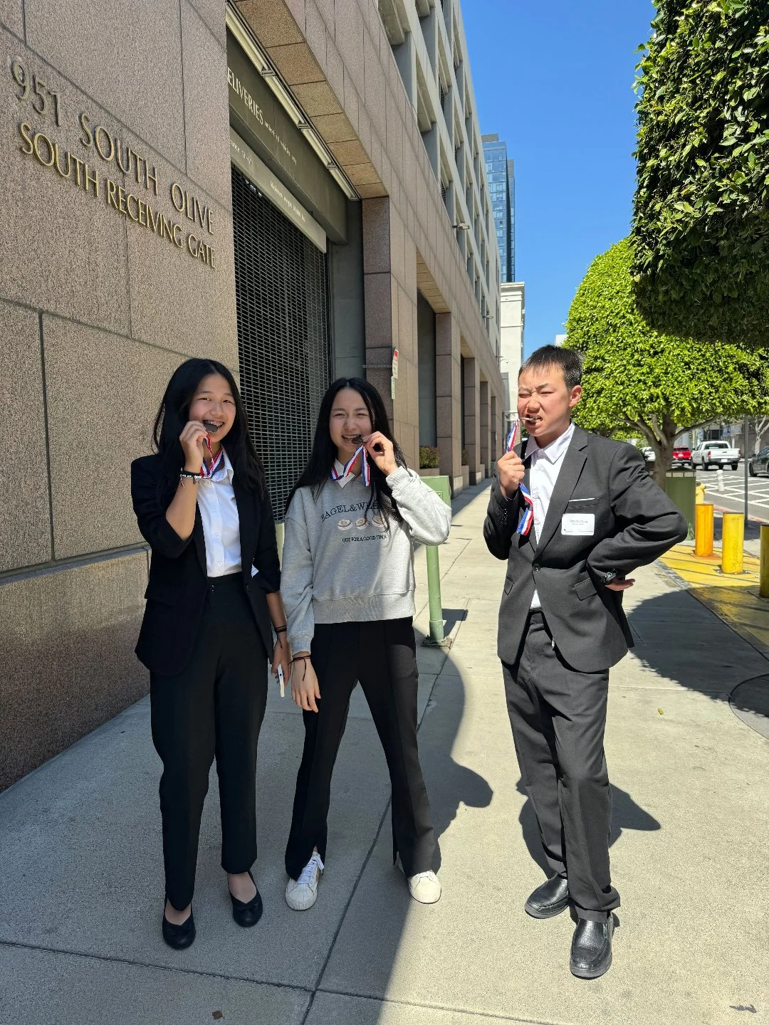 Three young people standing on a sidewalk outside a building, smiling and holding medals, with a clear blue sky and green trees in the background.