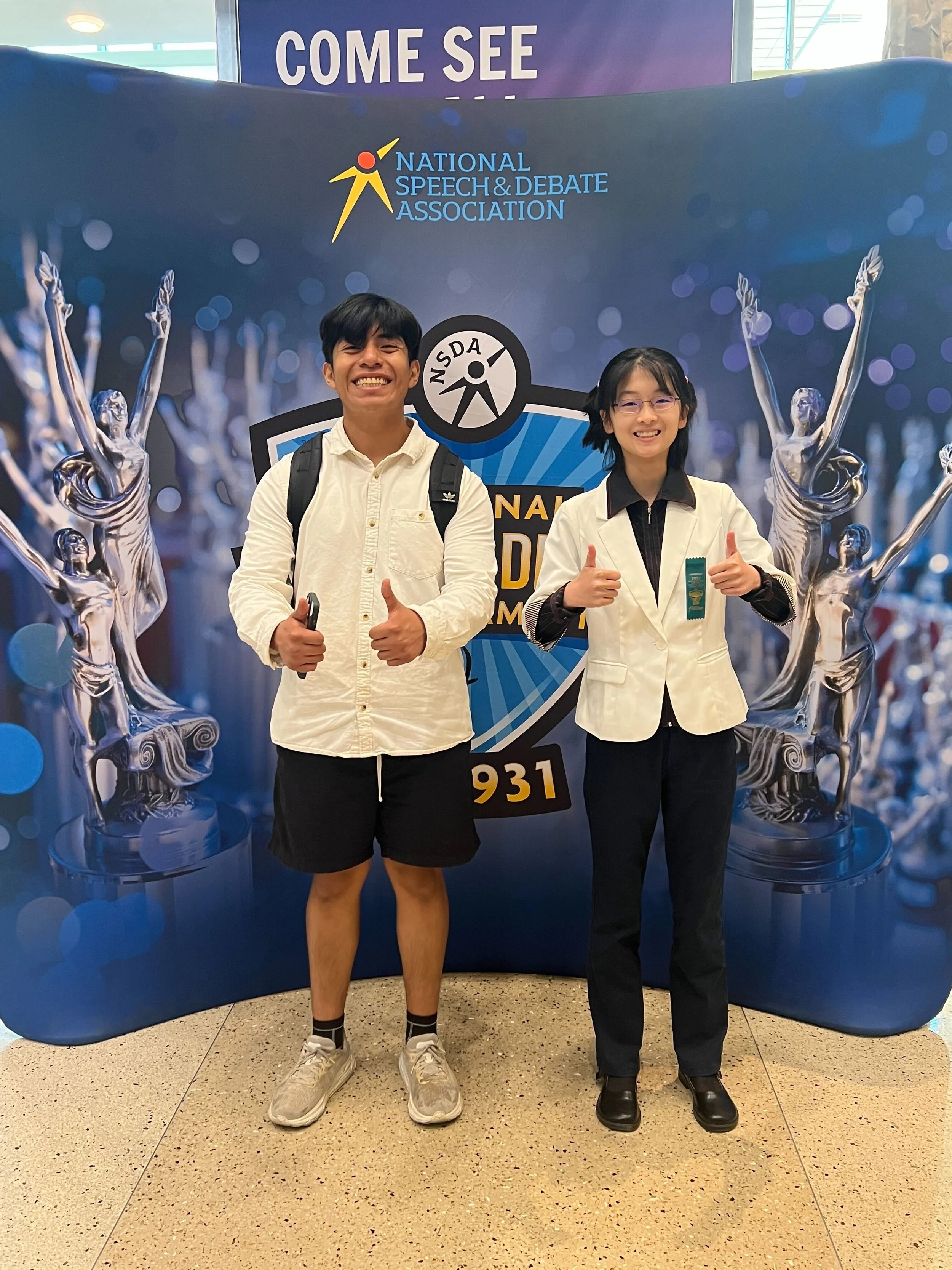 Two young people standing in front of a backdrop with the National Speech & Debate Association logo, giving thumbs up and smiling.