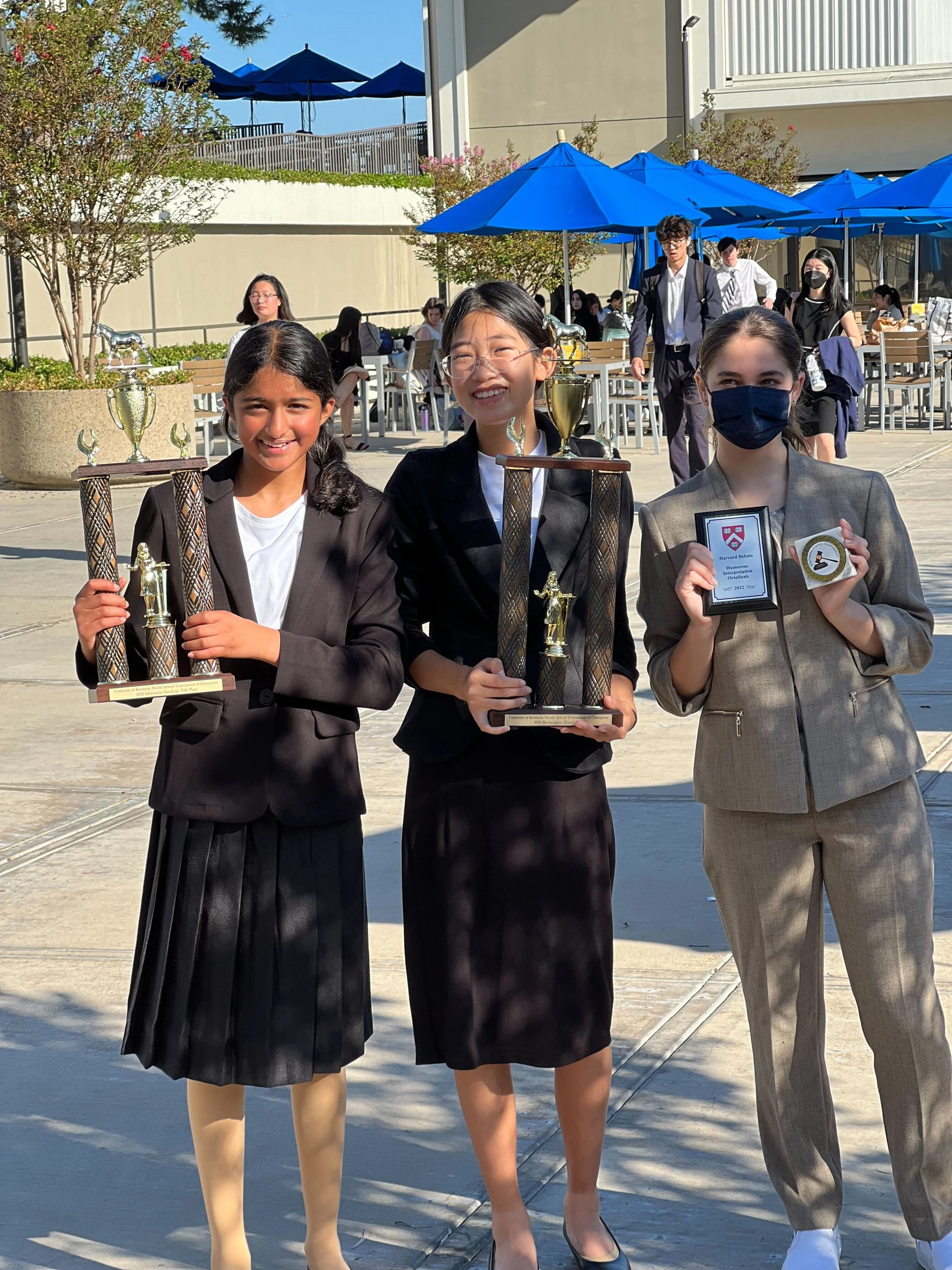 Three young girls in formal attire are standing outdoors, smiling, and holding trophies and awards, likely celebrating a recent achievement or competition.