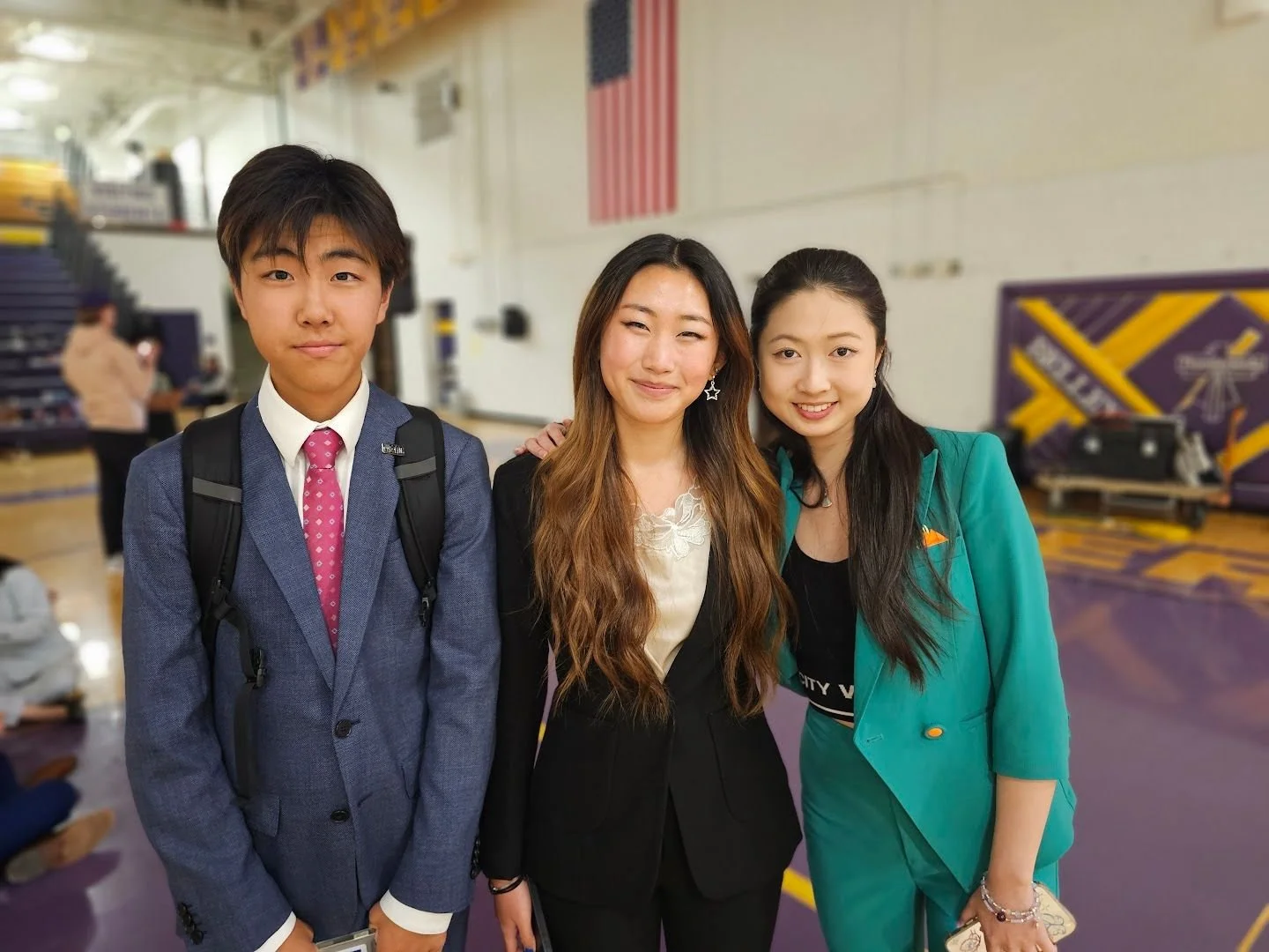 Three students in a gymnasium, smiling and posing for a photo. The boy on the left is wearing a blue suit, pink tie, and carrying a backpack. The woman in the middle has long, wavy hair, wearing a black blazer and cream top. The girl on the right has