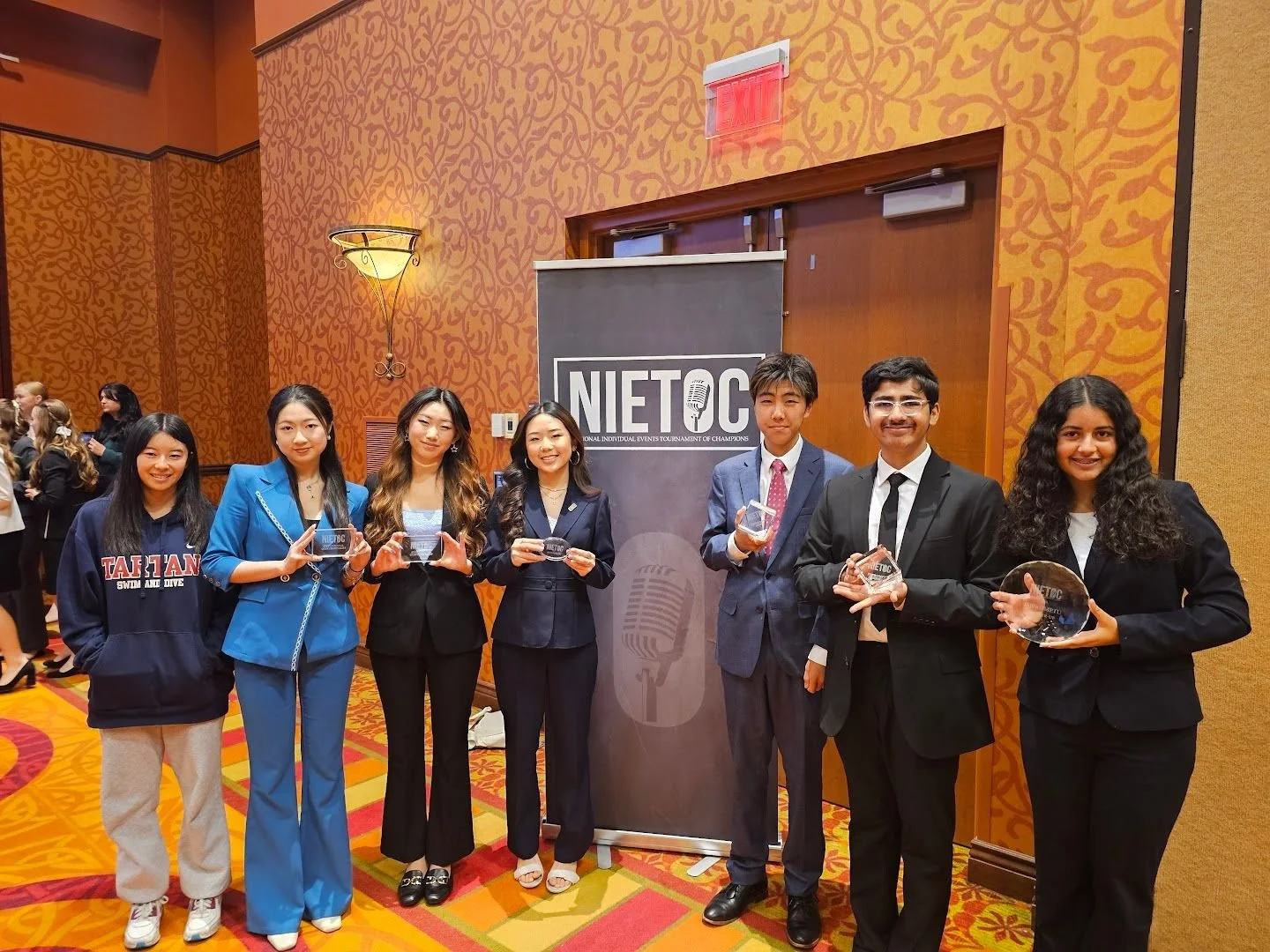 Group of seven young adults in formal and casual attire posing with awards at an event, standing in front of a banner with 'NIEC' logo, inside a warmly decorated conference room.