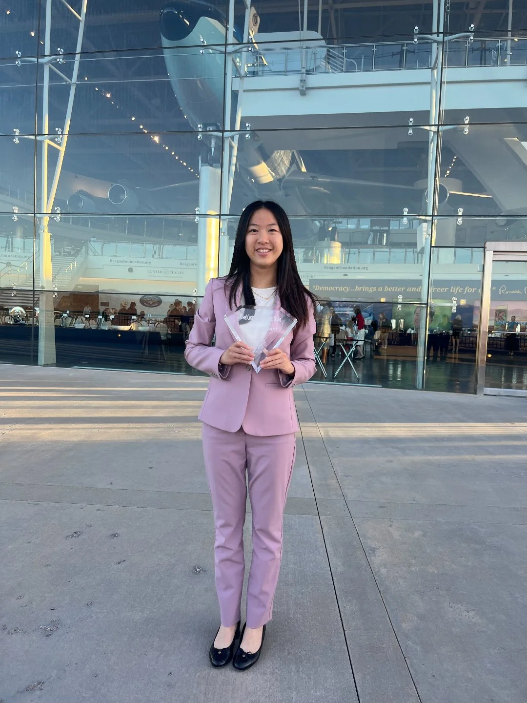 Woman in pink suit holding awards outside a modern glass building with an airplane visible inside.