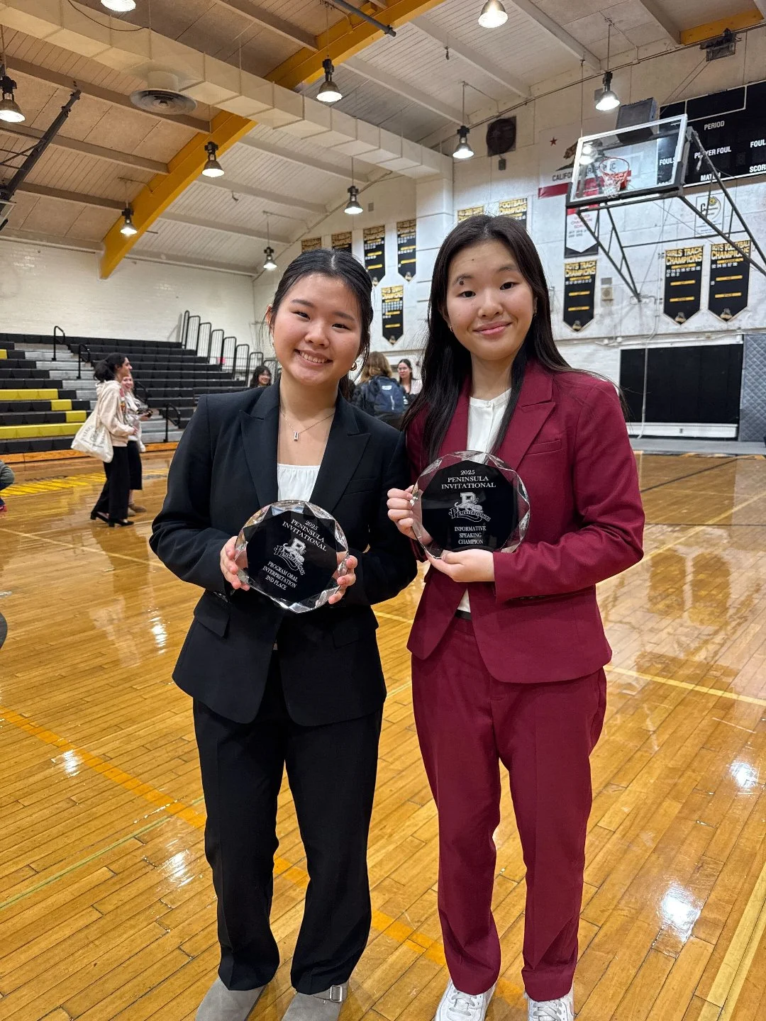 Two young women in business suits holding glass awards standing on a basketball court inside a gymnasium.