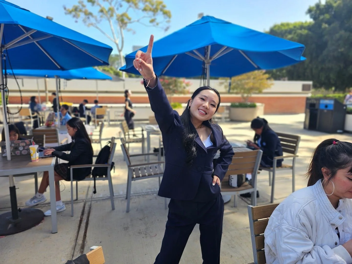 A woman in a navy blue blazer smiling and pointing upward with her right hand outdoors at a patio with several tables, chairs, and large blue umbrellas, with other people sitting and standing around.