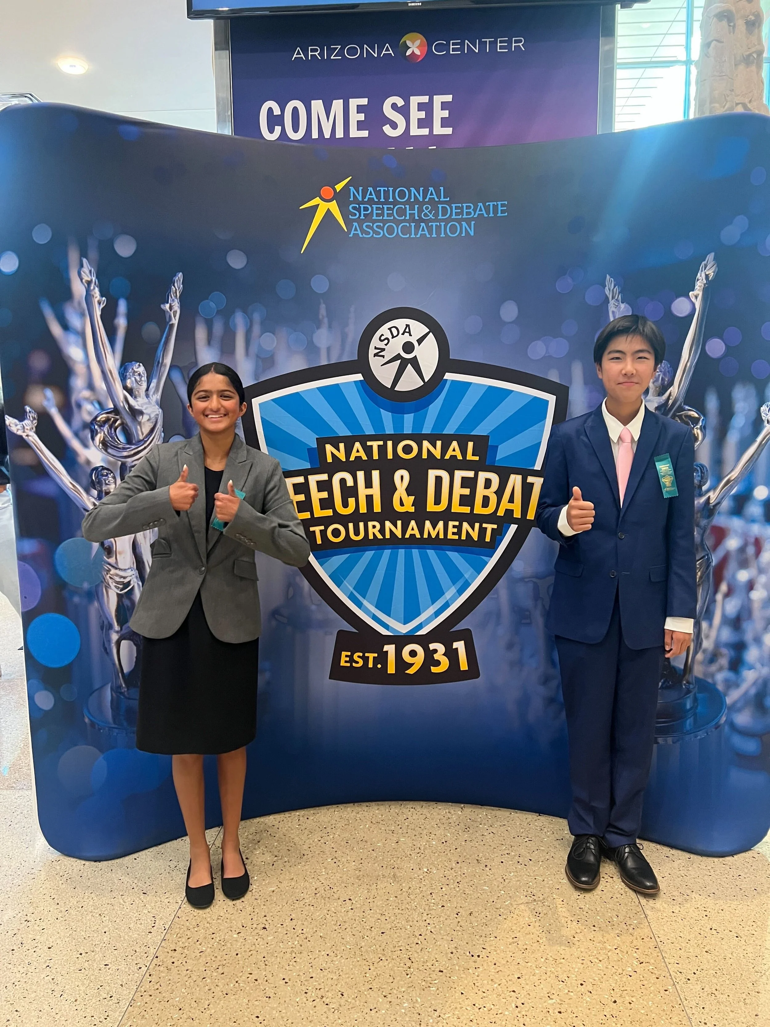 Two young students in professional attire standing in front of a promotional backdrop for the National Speech & Debate Tournament. The girl on the left is giving two thumbs up, wearing a gray blazer and black skirt, and the boy on the right is giving