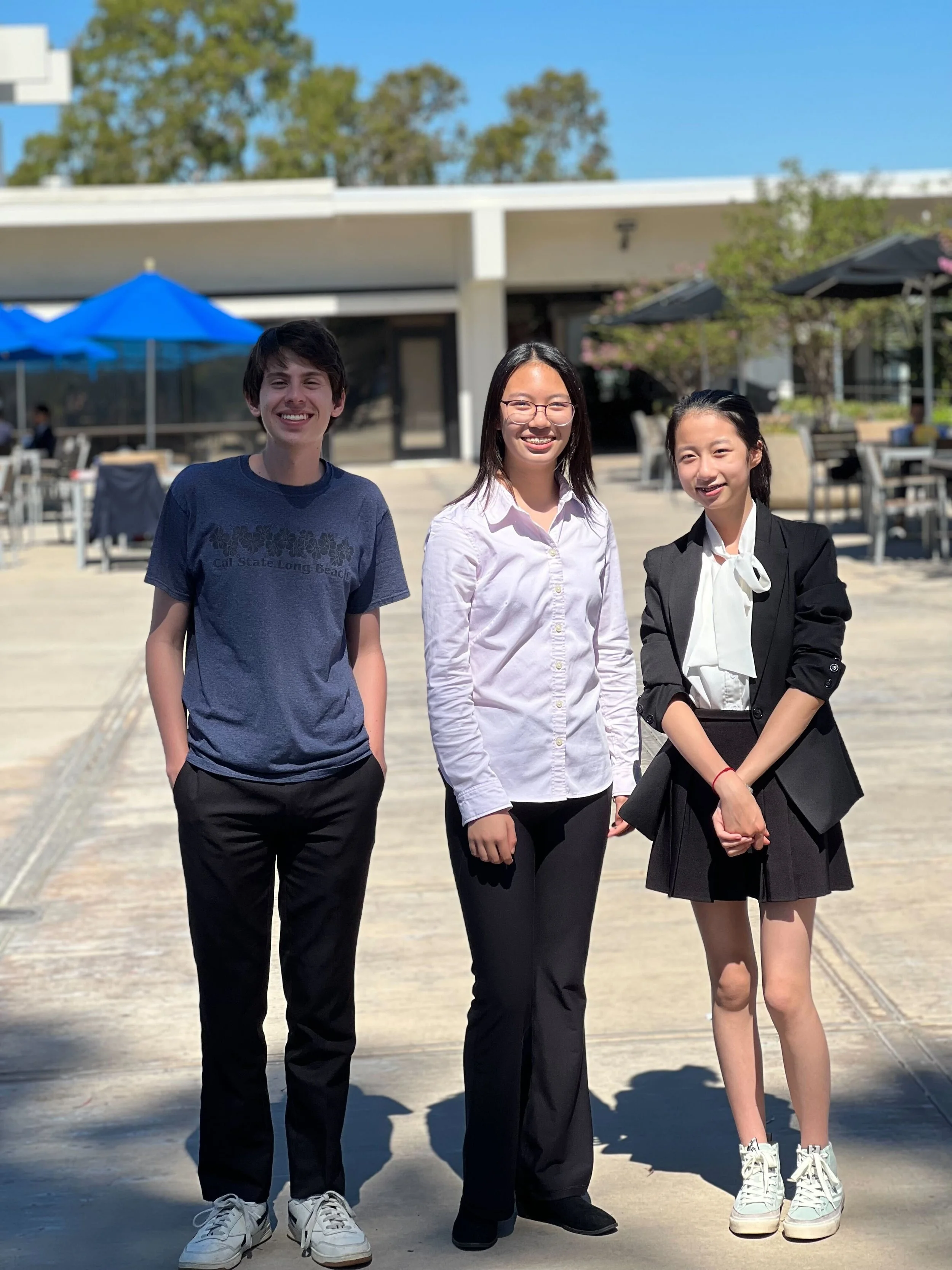 Three young people standing outdoors on a sunny day, smiling at the camera, with a building and outdoor seating area with umbrellas in the background.