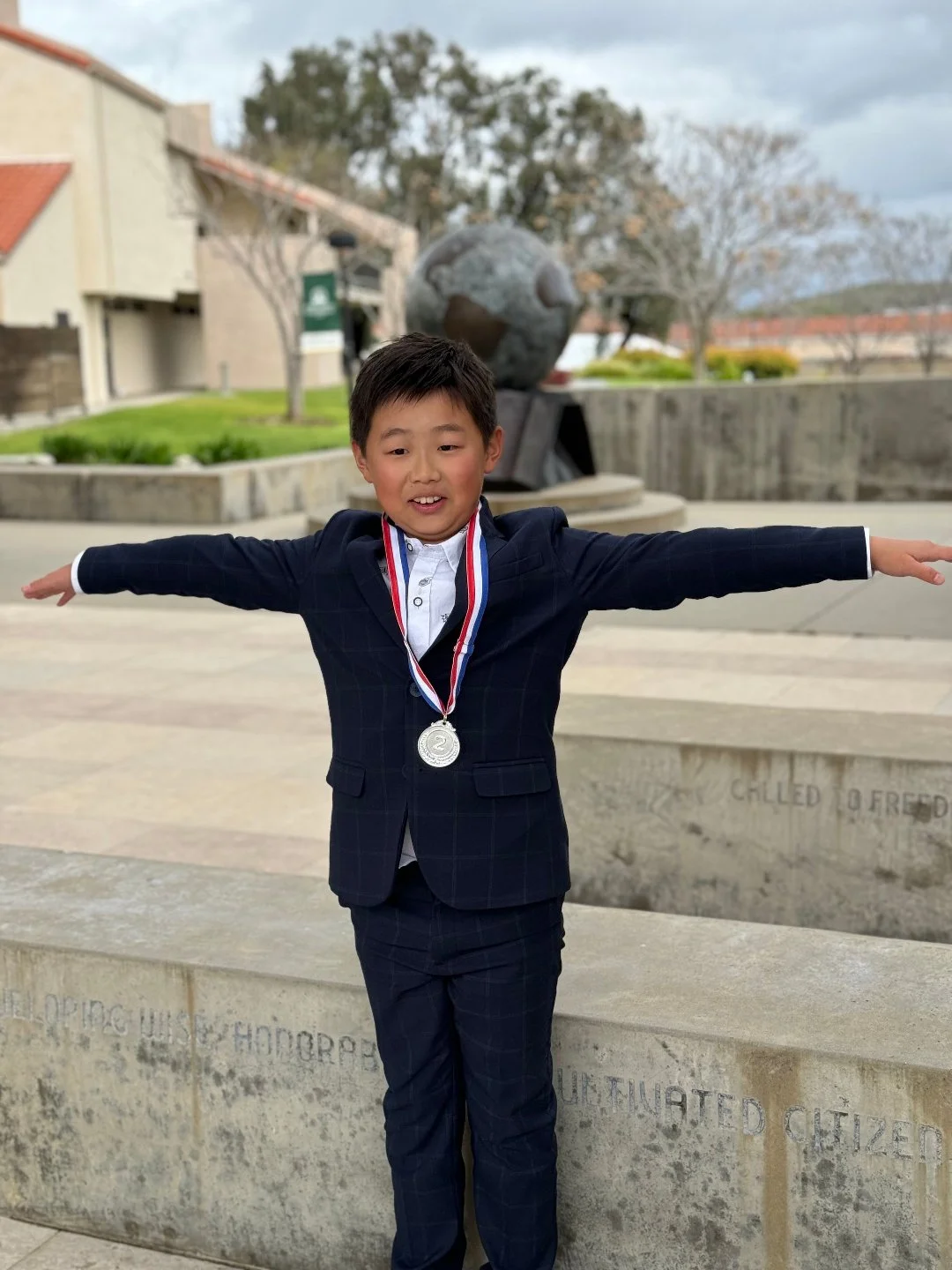 A young boy in a suit with a medal around his neck standing with arms outstretched in front of a memorial fountain. The boy is smiling, and the background shows trees, buildings, and a cloudy sky.