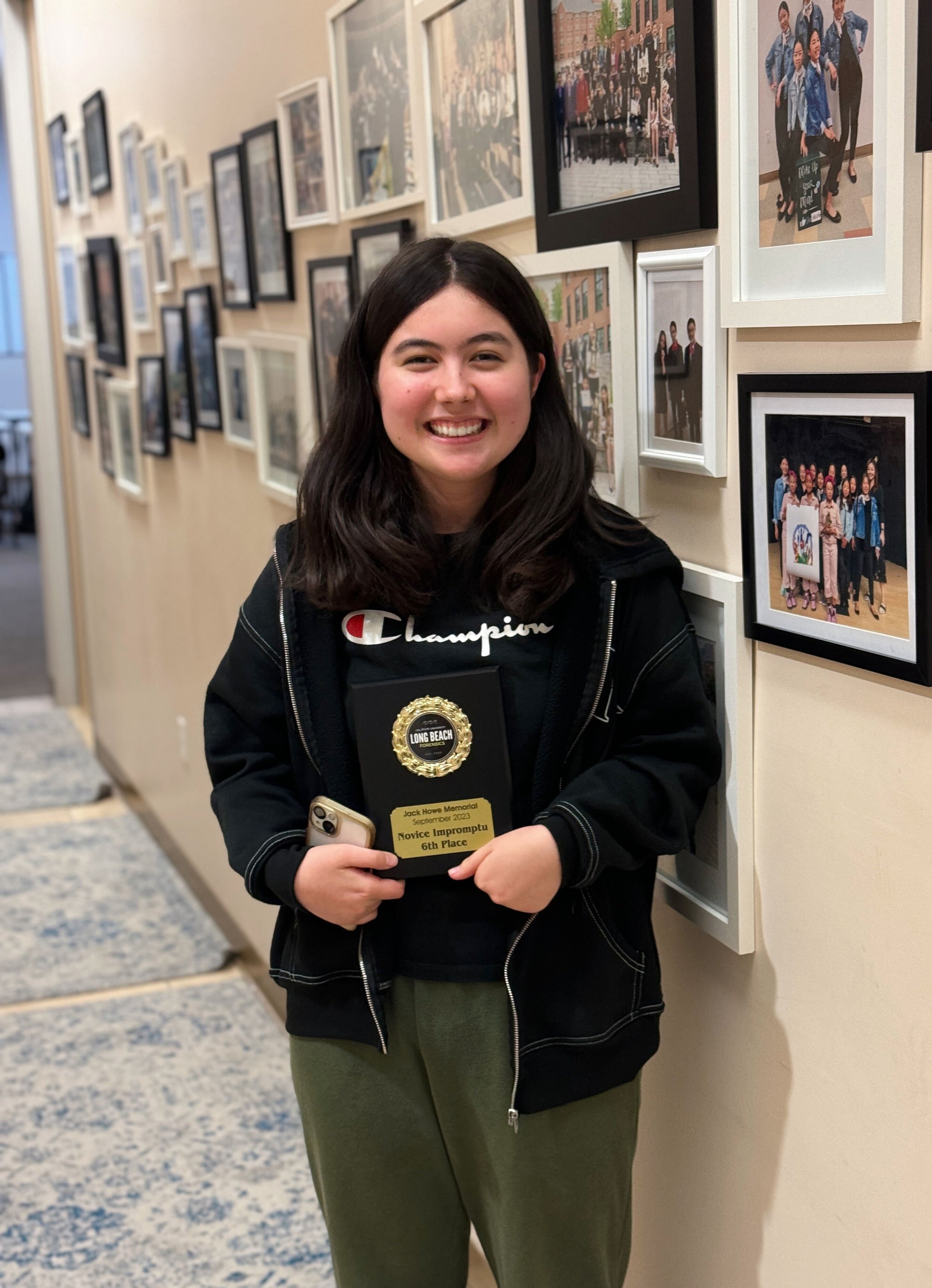 Young woman smiling and holding a plaque and phone, standing in front of a wall with framed photographs.