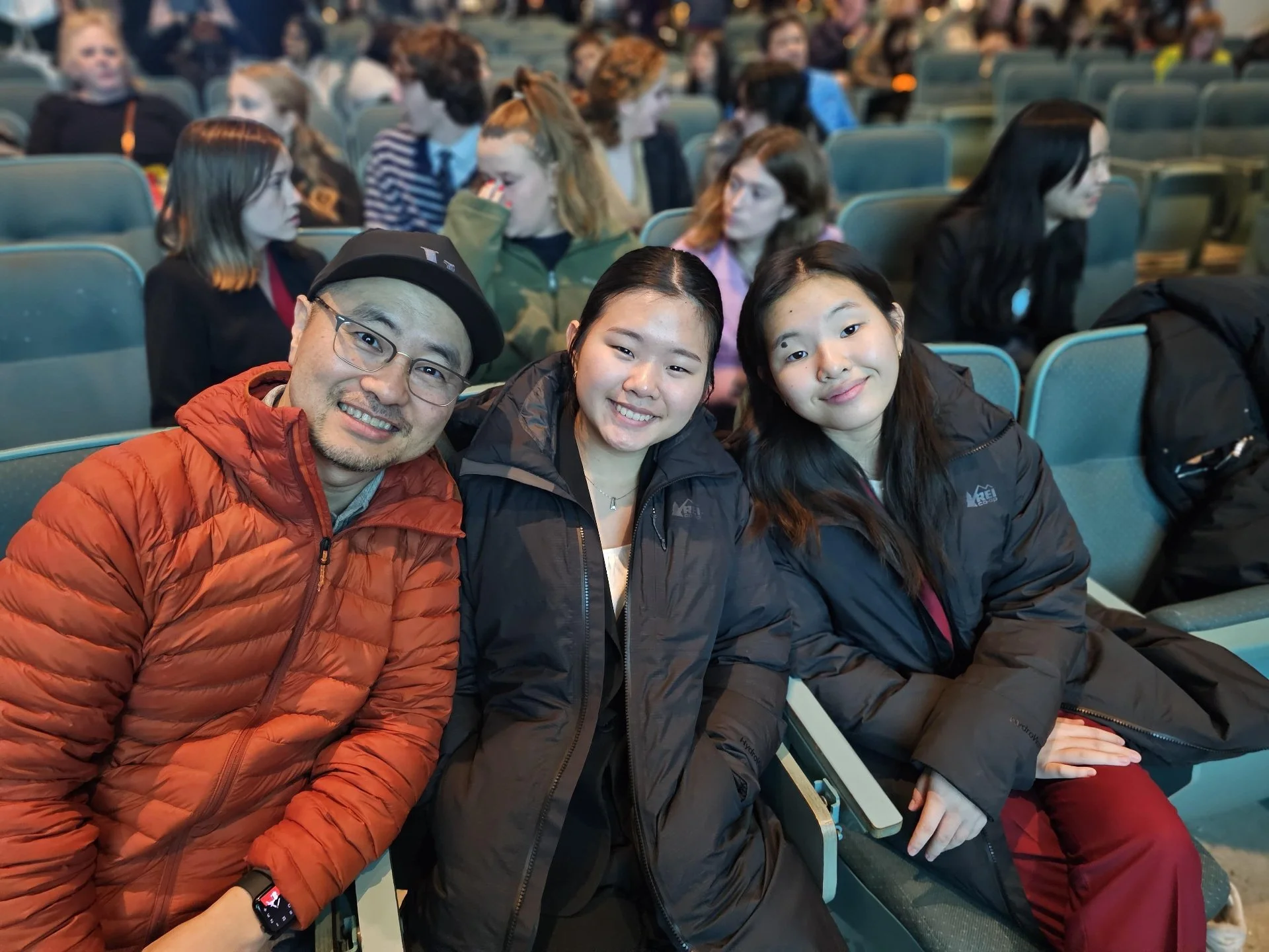 Three people sitting in an audience, smiling at the camera, with a crowd of people behind them.