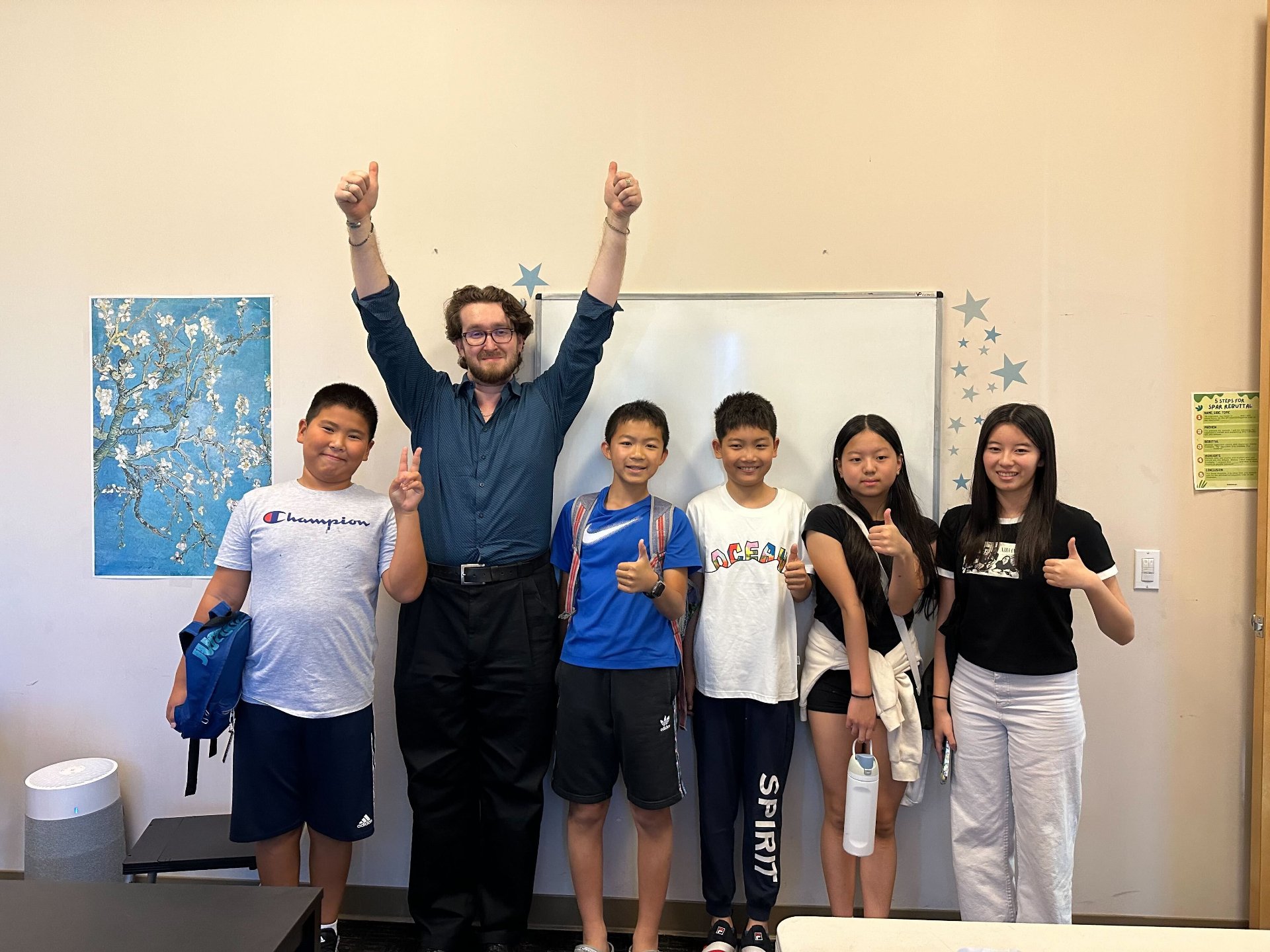 A diverse group of six children and one adult, all smiling and giving thumbs-up or peace signs, standing in a classroom in front of a whiteboard, with colorful wall decorations and a poster.