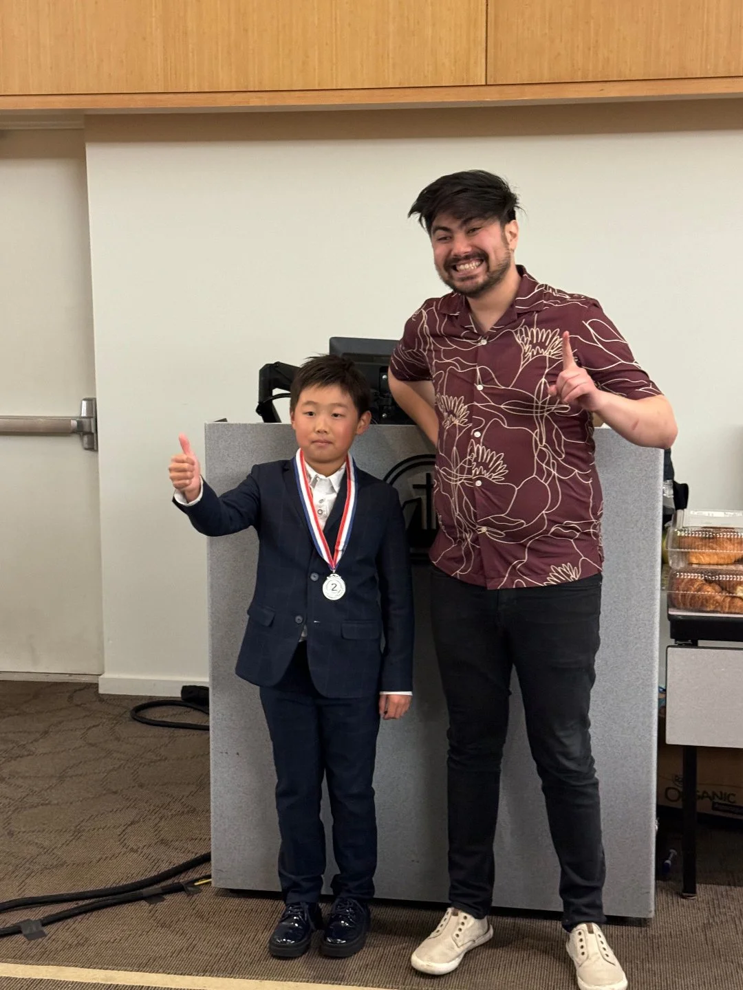 A young boy in a suit with a medal around his neck giving a thumbs-up, standing next to an adult man in casual clothing, also giving a thumbs-up, in a room with a grey podium and cake boxes in the background.
