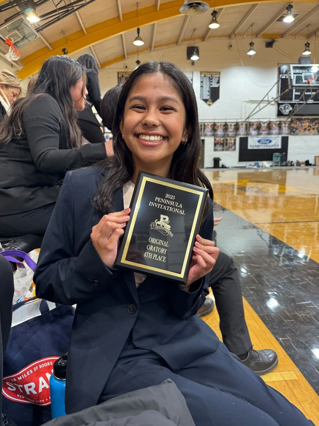 A young girl in a school uniform smiling and holding a plaque that reads "2025 Peninsula Invitational, Original Oratory, 4th Place," seated on a gymnasium floor surrounded by other students and spectators.