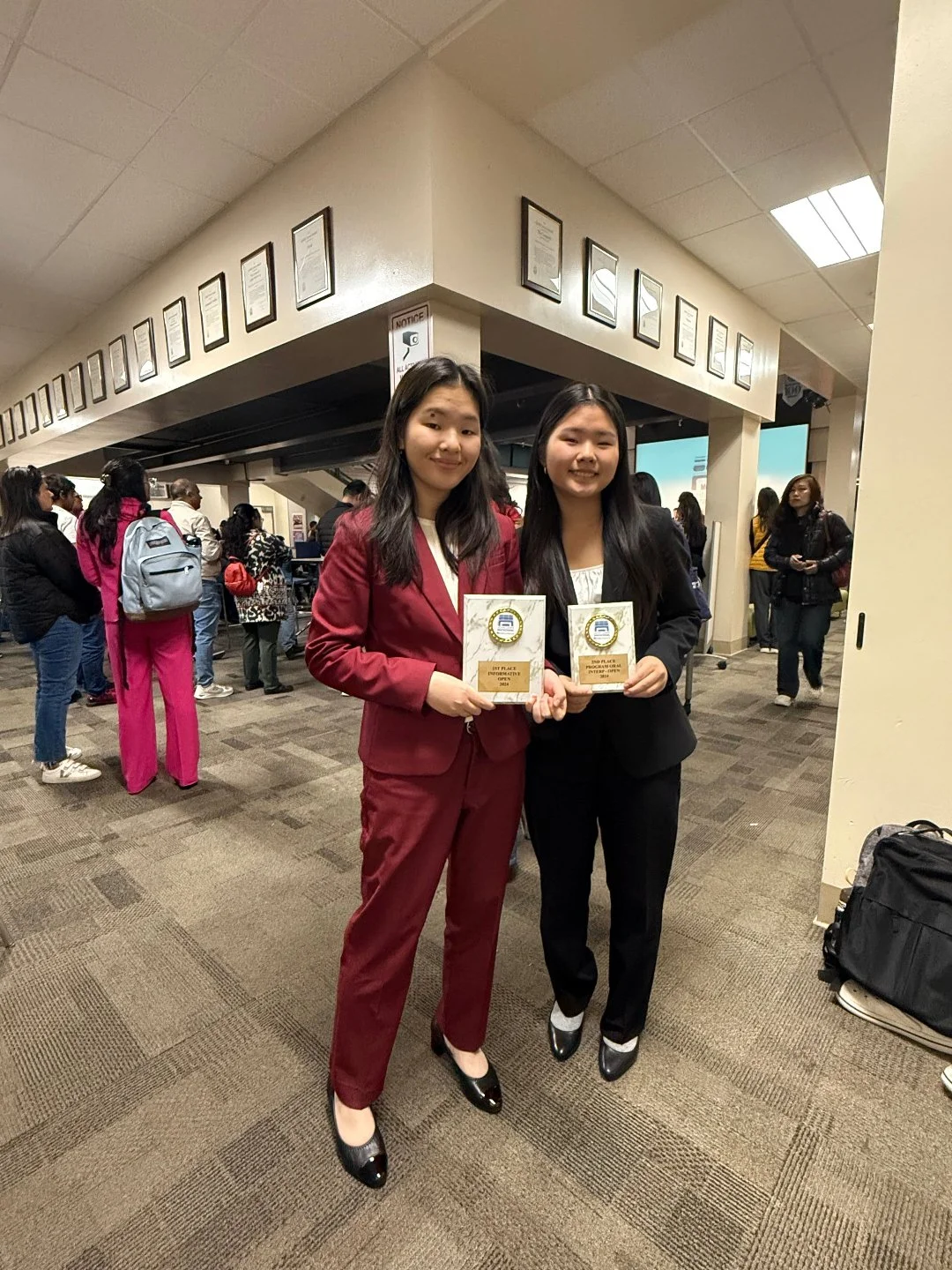 Two young women in business attire holding awards at an indoor event with other attendees in the background.