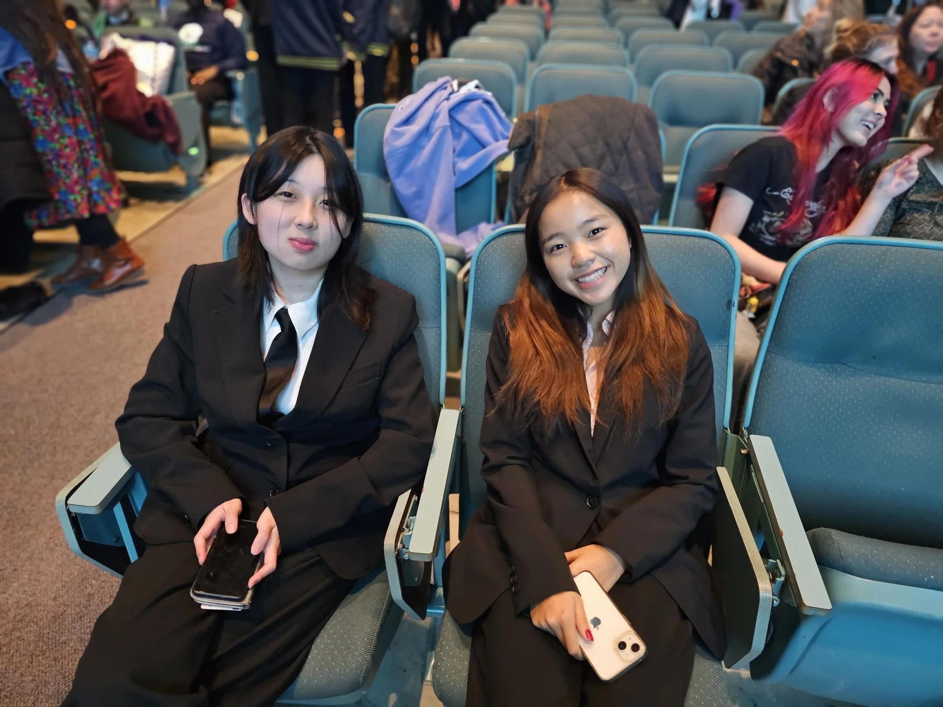Two young women in black suits and white shirts sitting in an auditorium, smiling and holding phones. One has black hair and the other has long brown hair. There are other people in the background, some chatting and some standing.