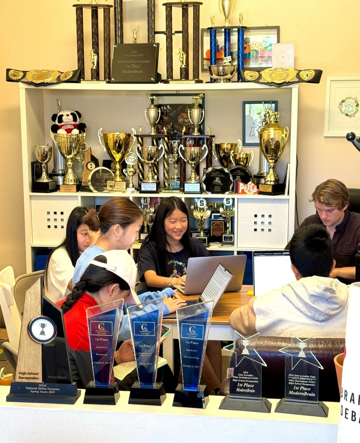 Group of young students working on laptops in a room filled with trophies and awards, with a shelf of plaques and trophies in the background.