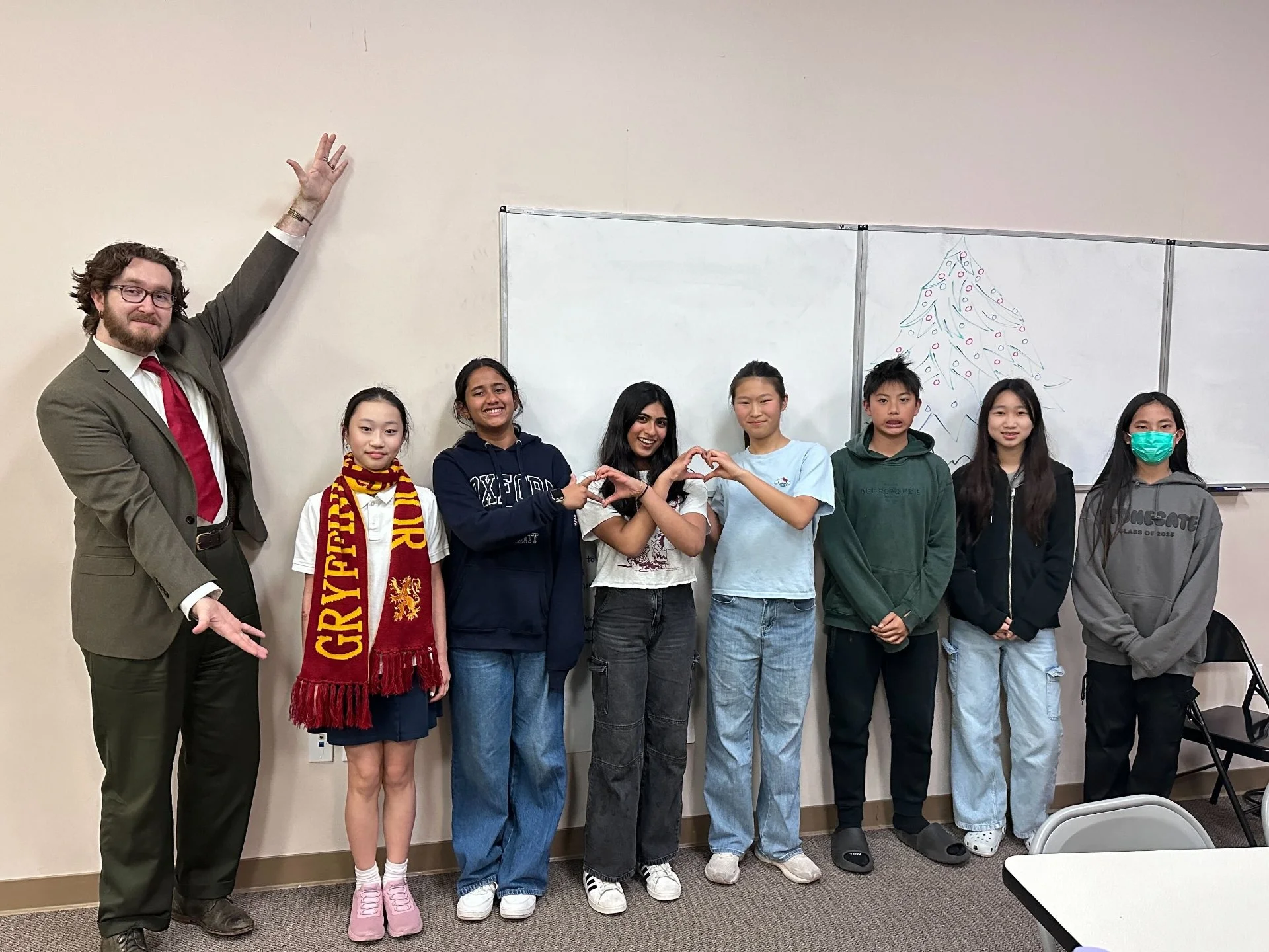 Group of eight people, including a man in a beige suit with a red tie and seven students, standing in front of a whiteboard with a holiday-themed drawing of a Christmas tree, inside a classroom.