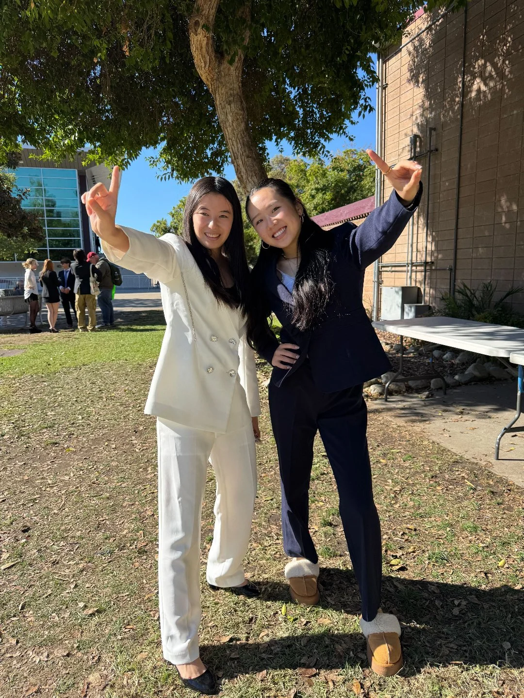 Two young women dressed in formal attire, smiling and posing outdoors under a tree on a sunny day, with a group of people standing in the background.