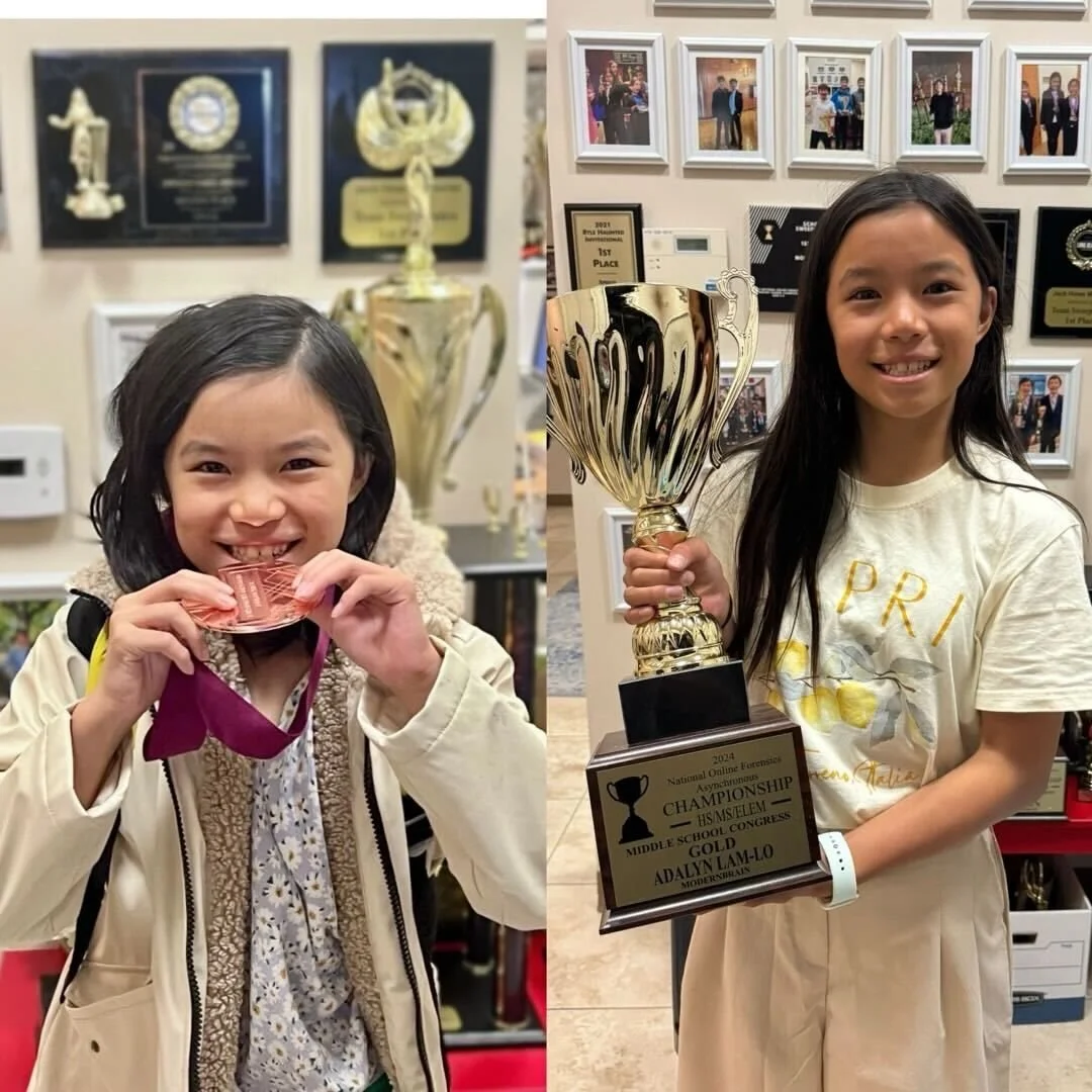 Two young girls, one holding a medal and the other holding a large trophy, smiling indoors with award plaques and framed photos on the walls behind them.